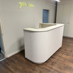 LookMeble - Biała lada recepcyjna z ryflowanym frontem na tle drewnianej podłogi i białych szaf wnękowych. Nad ladą neonowy napis 'you can'.