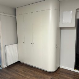 LookMeble - Biała szafa na wymiar z zaokrąglonym bokiem, dopasowana do wnętrza. Cztery fronty z małymi, okrągłymi uchwytami. Ciemna podłoga, jasne ściany. Widoczny grzejnik.