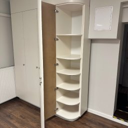 LookMeble - Biała szafa wnękowa z półkami narożnymi, otwarte drzwi ukazują wnętrze. Widoczna podłoga z paneli i fragment ściany z drzwiami. Zaokrąglone krawędzie szafy.