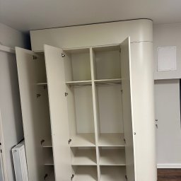 LookMeble - Biała szafa wnękowa z otwartymi drzwiami, widoczne półki i drążki na ubrania. Zaokrąglony bok szafy, minimalistyczny design, drewniana podłoga.