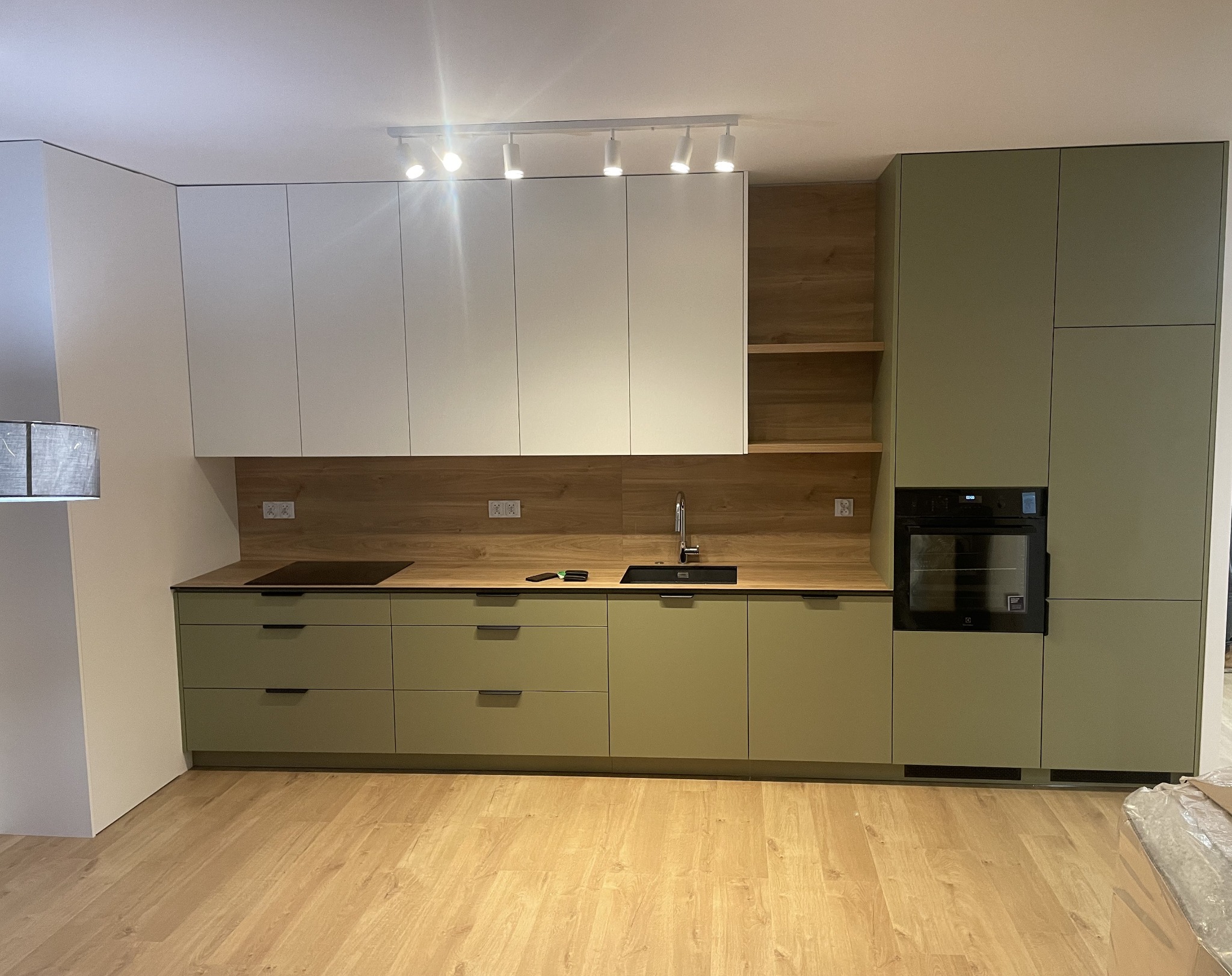 Nowoczesna kuchnia na wymiar w odcieniach zieleni i bieli, z drewnianym blatem i oświetleniem punktowym. Widoczny piekarnik i płyta indukcyjna.