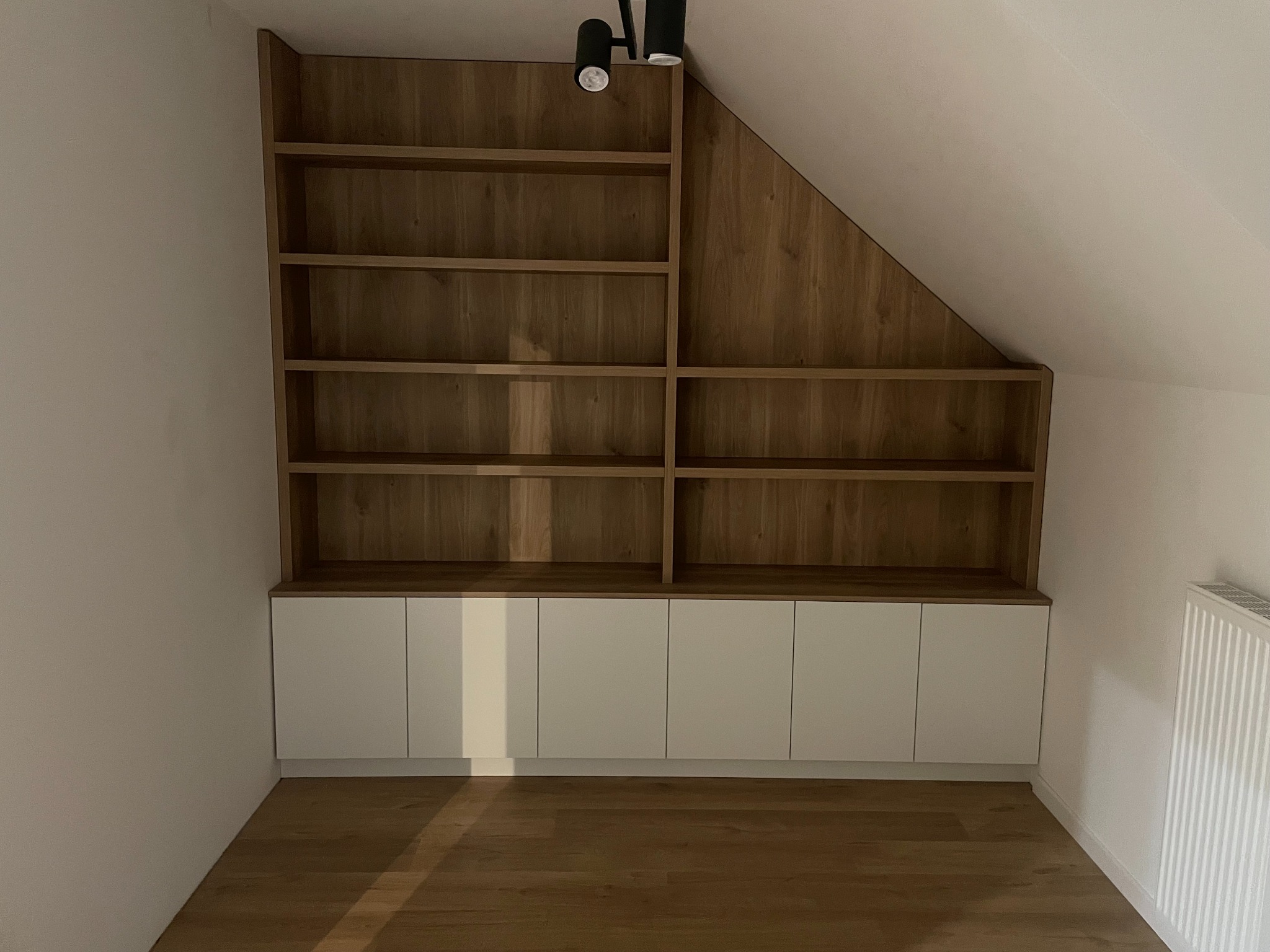Drewniana zabudowa pod skosem z białymi szafkami na dole, dopasowana do przestrzeni. Nad nią lampa sufitowa. Podłoga z paneli.