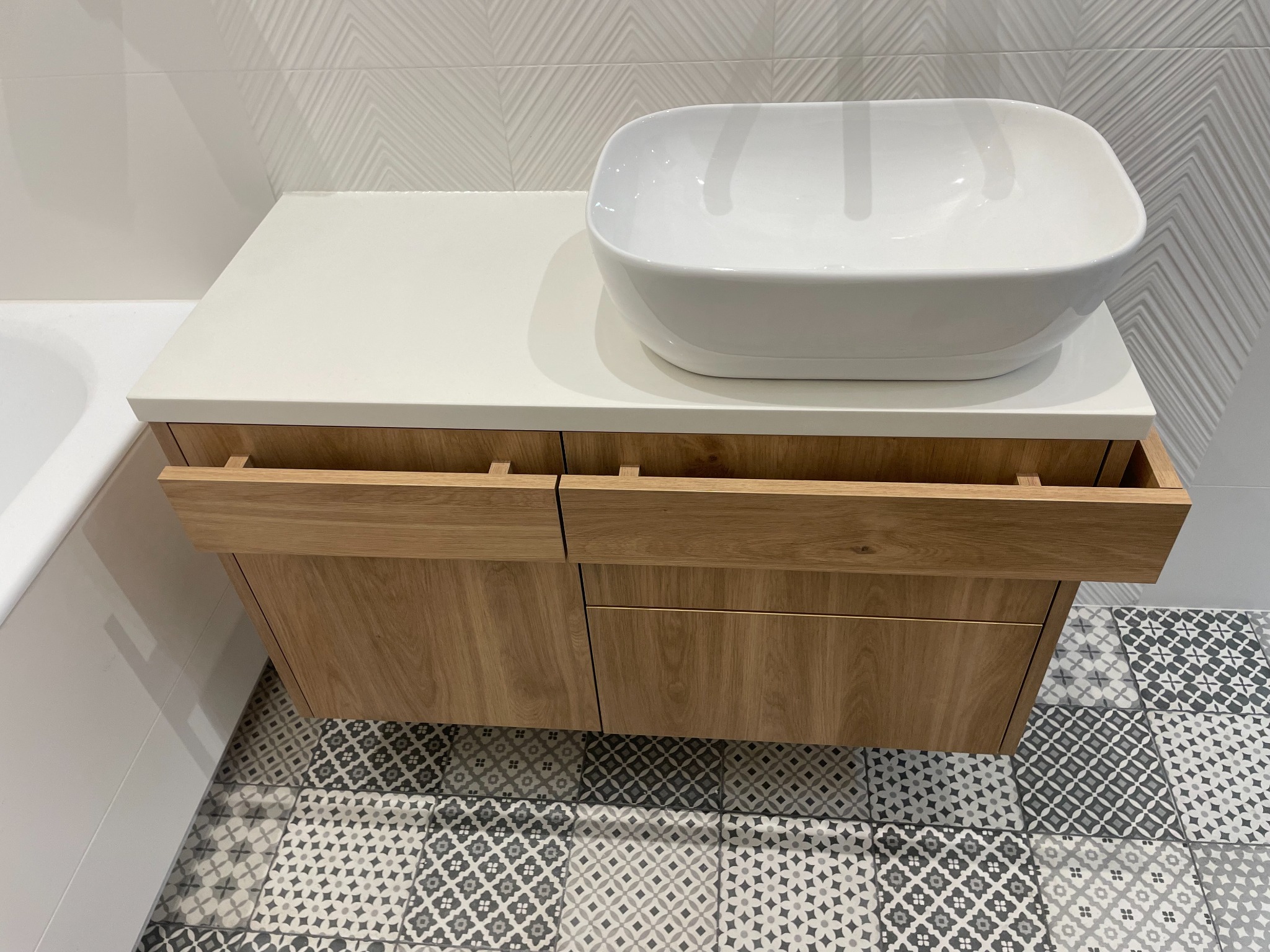 Drewniana szafka łazienkowa z blatem i umywalką nablatową. Widoczne otwarte szuflady i wzorzysta podłoga. Minimalistyczny design.