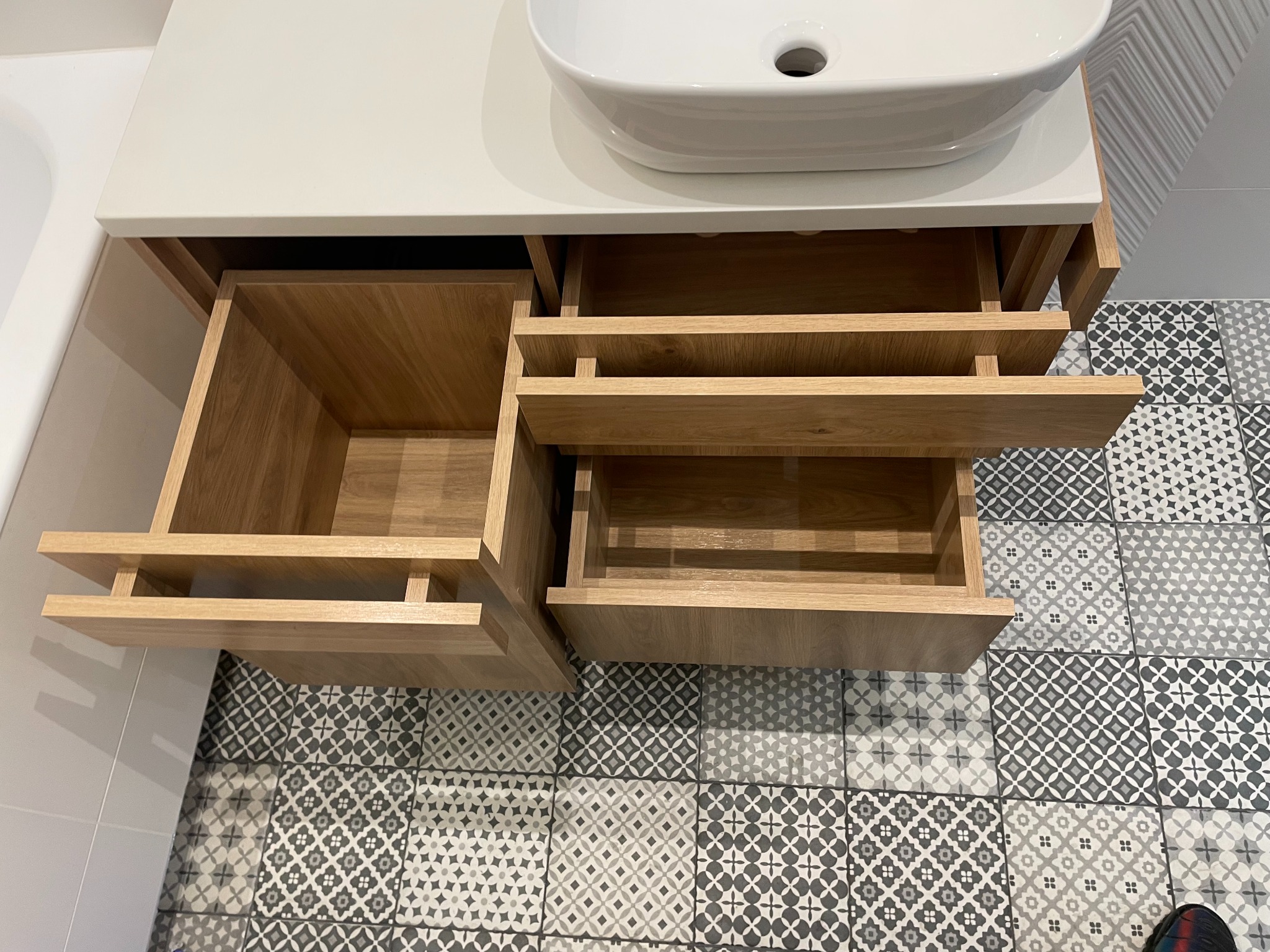 Szafka łazienkowa z umywalką i otwartymi szufladami z jasnego drewna na tle wzorzystej podłogi w szarościach. Widok z góry.
