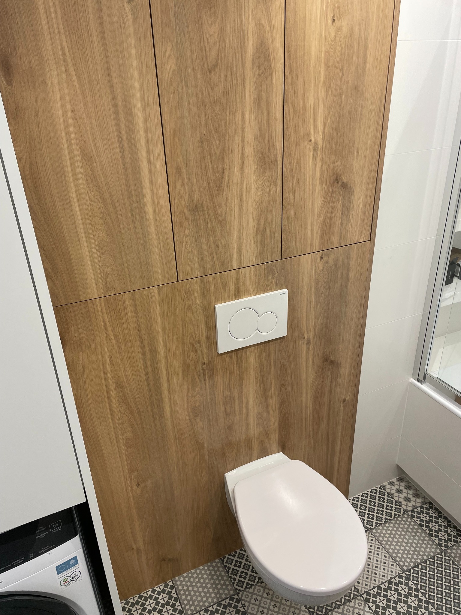 Drewniana szafka na wymiar w łazience, zabudowa toalety, biała armatura, wzorzysta podłoga. Naturalne oświetlenie, minimalistyczny design.