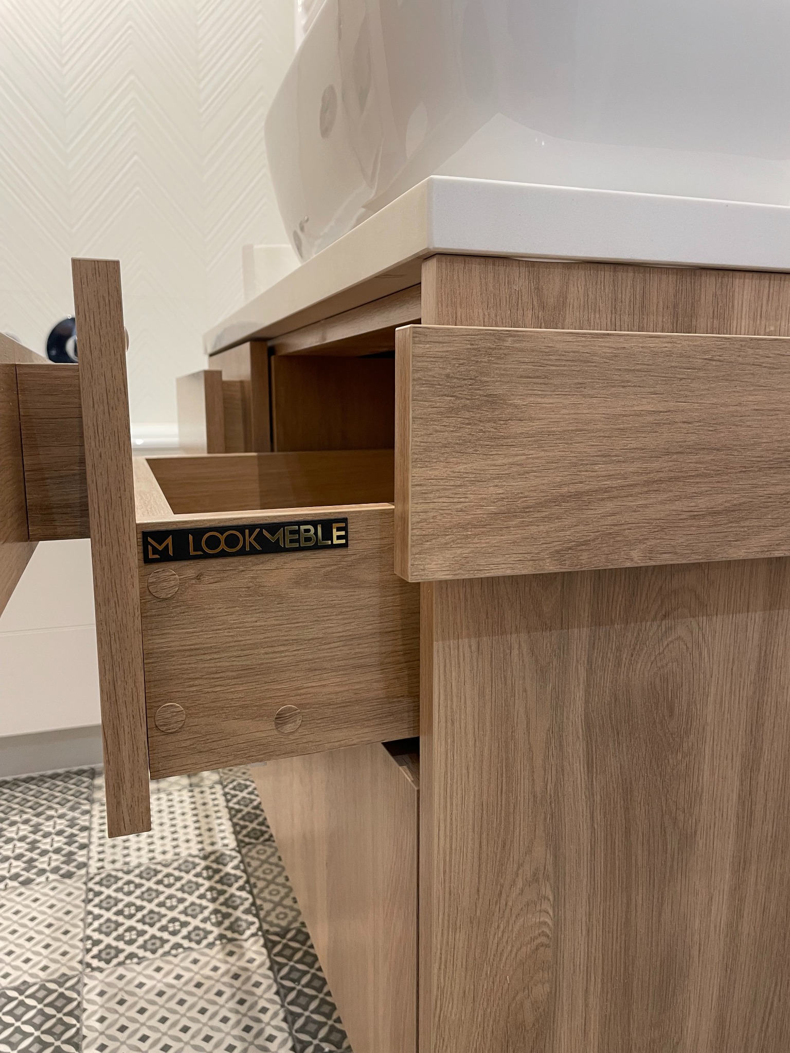 Otwarta szuflada szafki łazienkowej z jasnego drewna, widoczne logo 'M LOOKMEBLE', biała umywalka nablatowa, wzorzysta podłoga. Detal zabudowy meblowej.