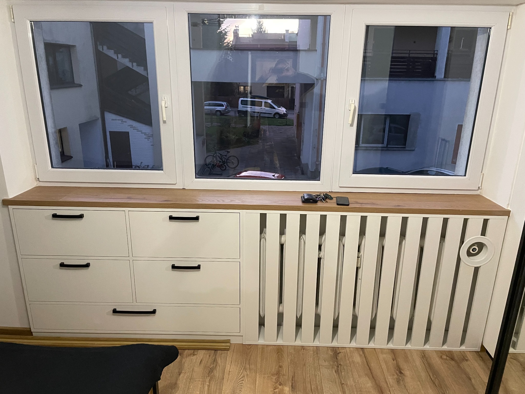 Zabudowa okna z białymi szafkami z czarnymi uchwytami i dekoracyjnym grzejnikiem. Blat drewniany, widok na podwórko z rowerem i samochodami.
