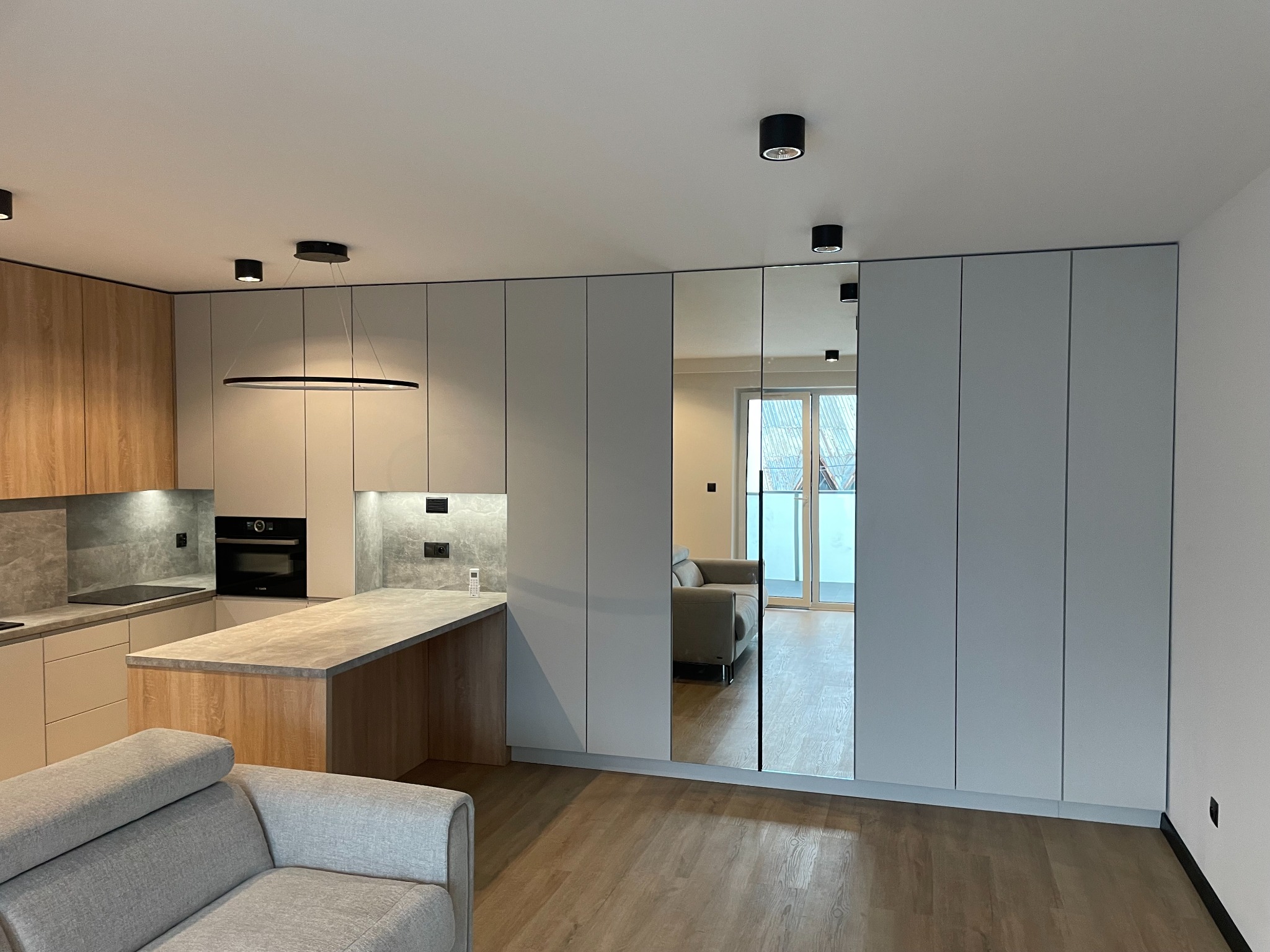 Nowoczesna kuchnia z wyspą i szafą wnękową z lustrzanymi drzwiami, widok na salon. Jasne drewno i szarość tworzą spójną całość.