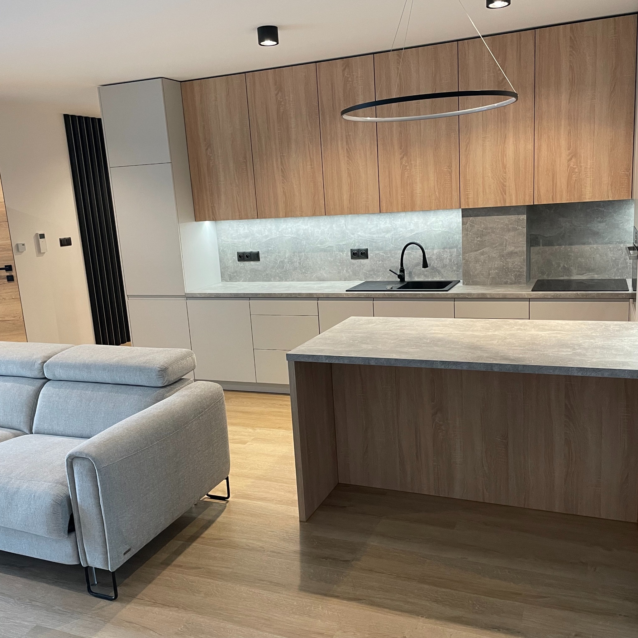 Nowoczesna kuchnia z drewnianymi frontami i wyspą, oświetlona lampą pierścieniową. Szary narożnik w tle. Minimalistyczny design z akcentami czerni.