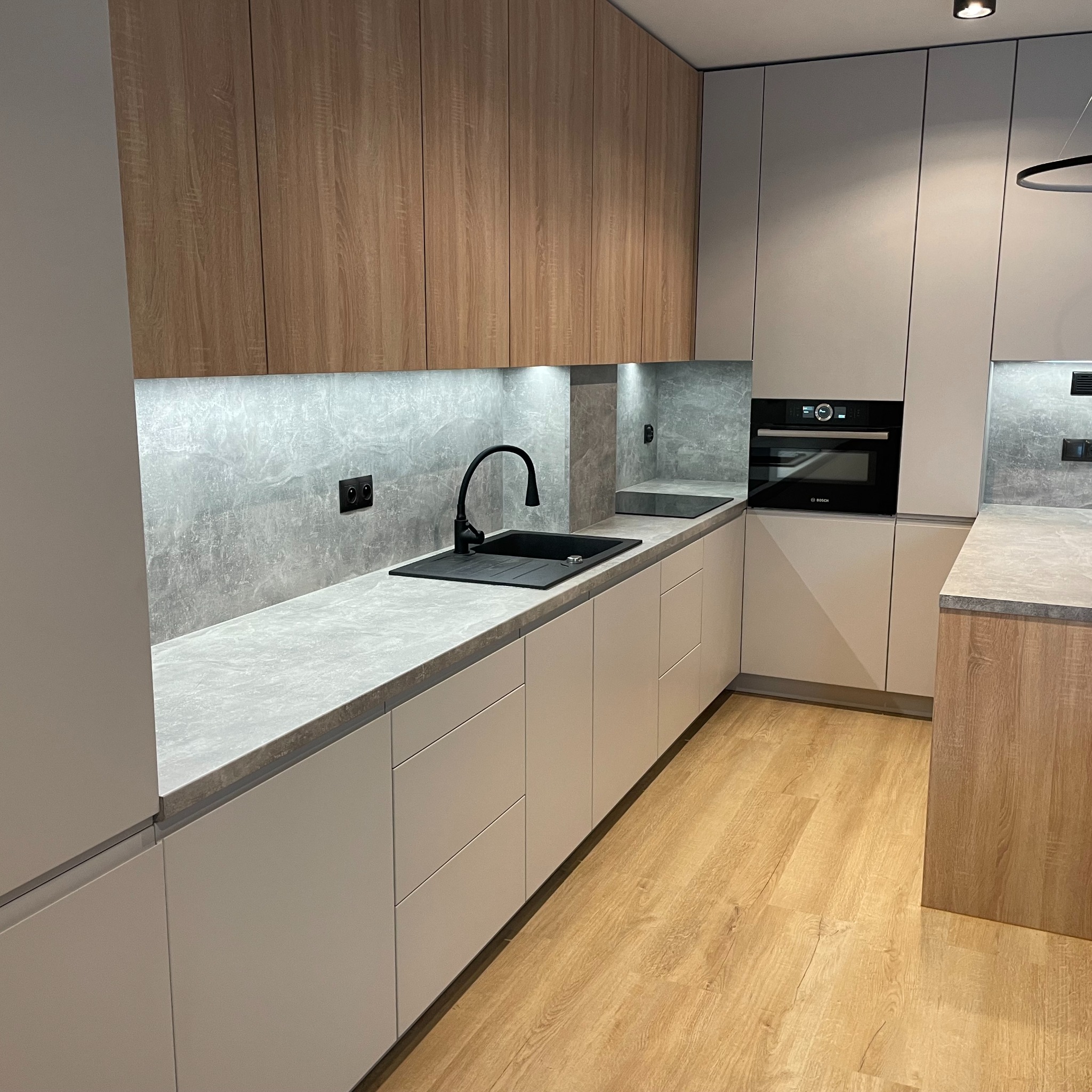 Nowoczesna kuchnia z drewnianymi i szarymi frontami, czarną armaturą i blatem imitującym kamień. Oświetlenie LED podszafkowe podkreśla detale wykończenia.