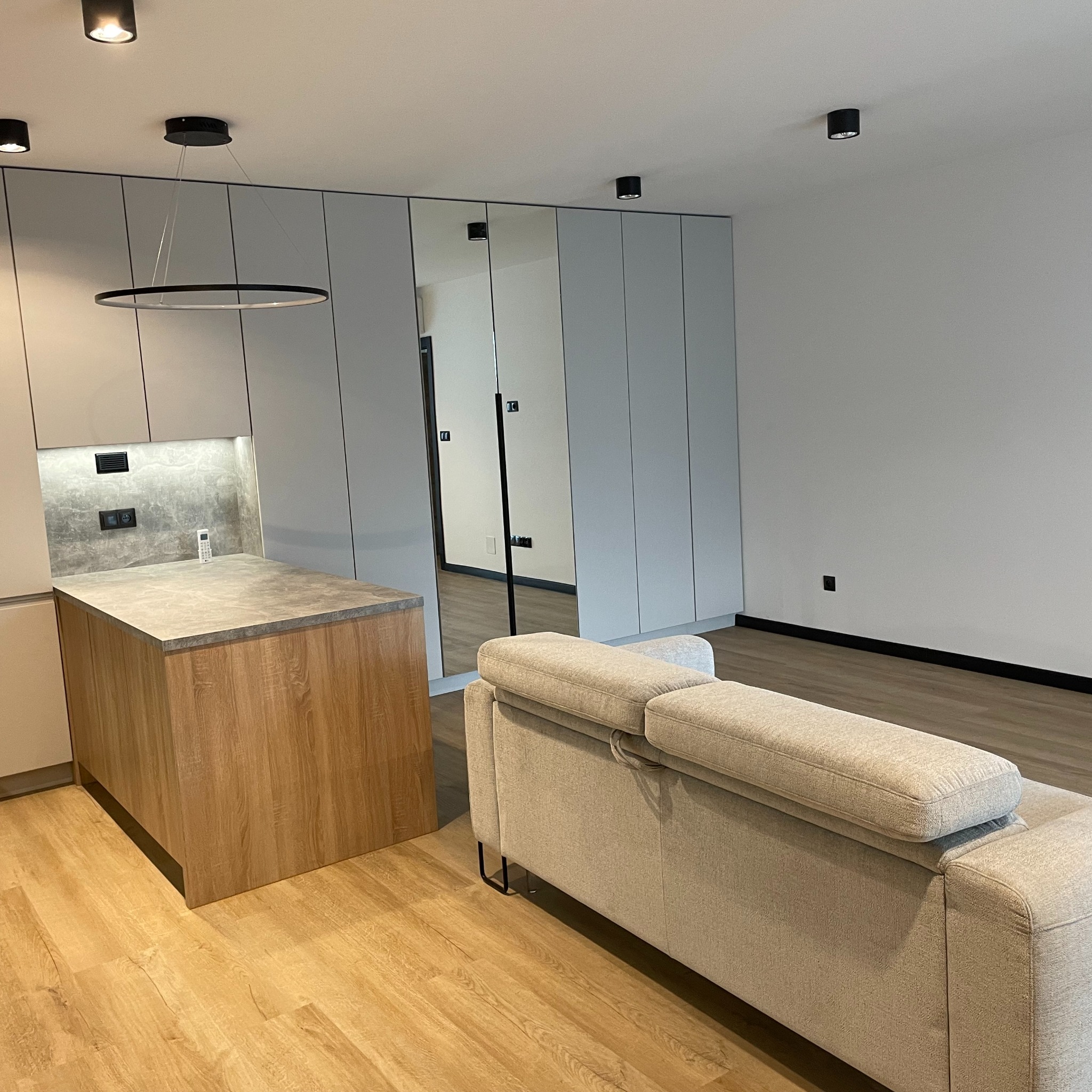 Nowoczesne wnętrze: szara zabudowa szafy z lustrami, drewniana wyspa kuchenna i beżowa sofa. Minimalistyczny design z okrągłą lampą sufitową.