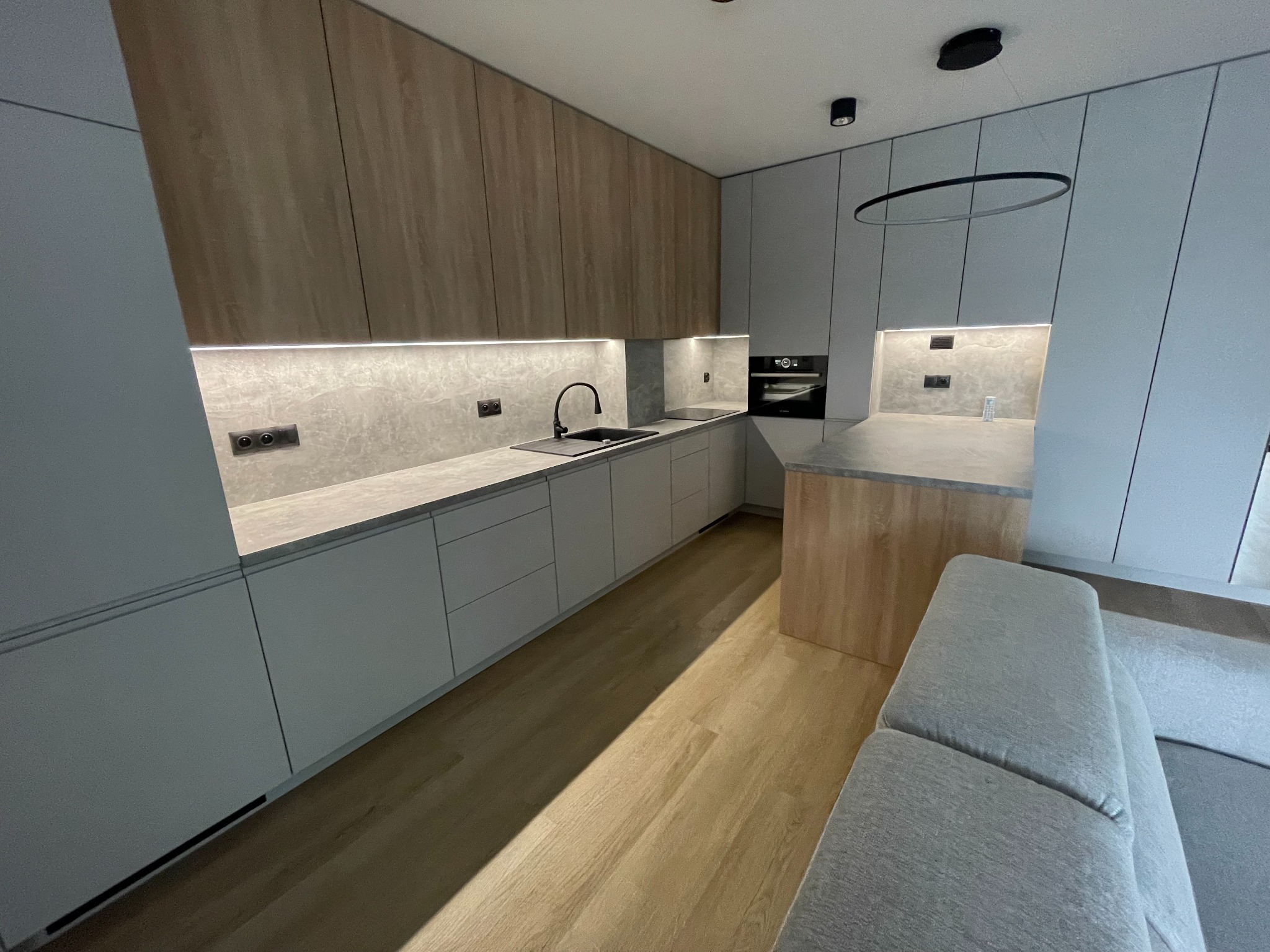 Nowoczesna kuchnia z drewnianymi i szarymi frontami, czarną armaturą i okrągłą lampą sufitową. Minimalistyczny design z oświetleniem LED pod szafkami.