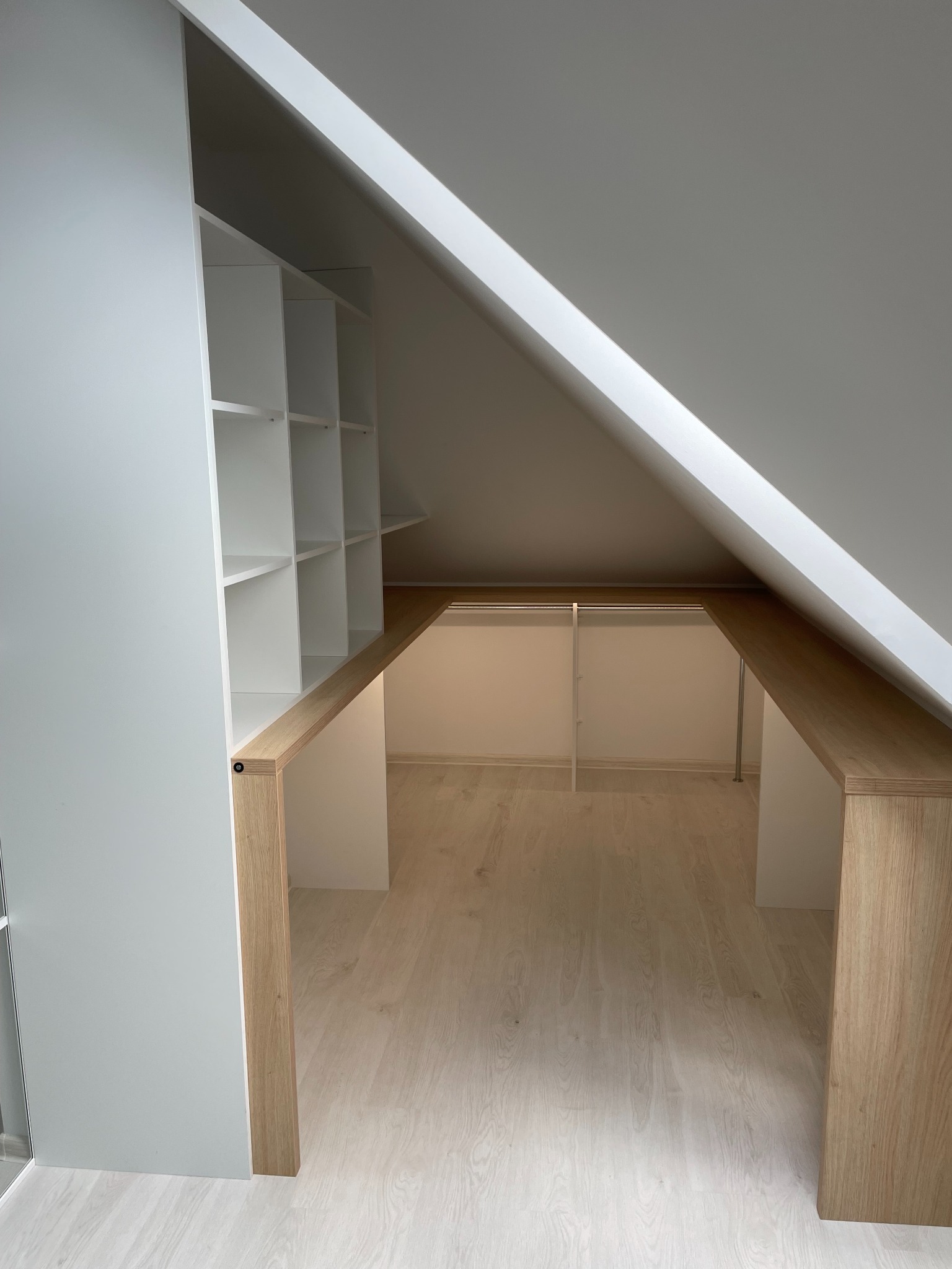 Funkcjonalna, otwarta garderoba w skosach poddasza z białymi półkami i drewnianymi elementami wykończeniowymi. Oświetlenie wewnątrz.