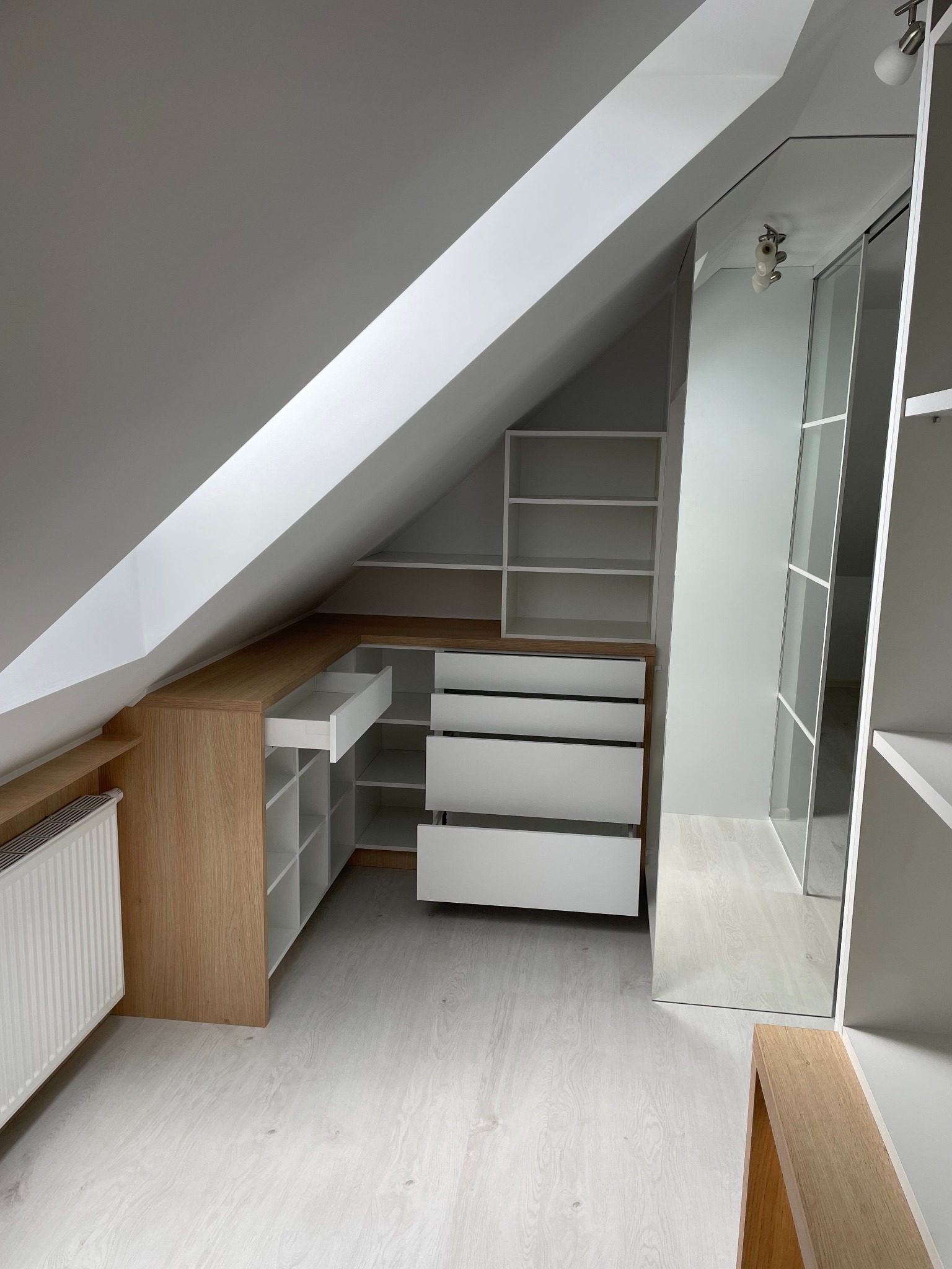 Szafa wnękowa na poddaszu z drewnianymi elementami i białymi frontami. Otwarta szuflada, lustrzane drzwi przesuwne, białe półki. Widok na skośny sufit.