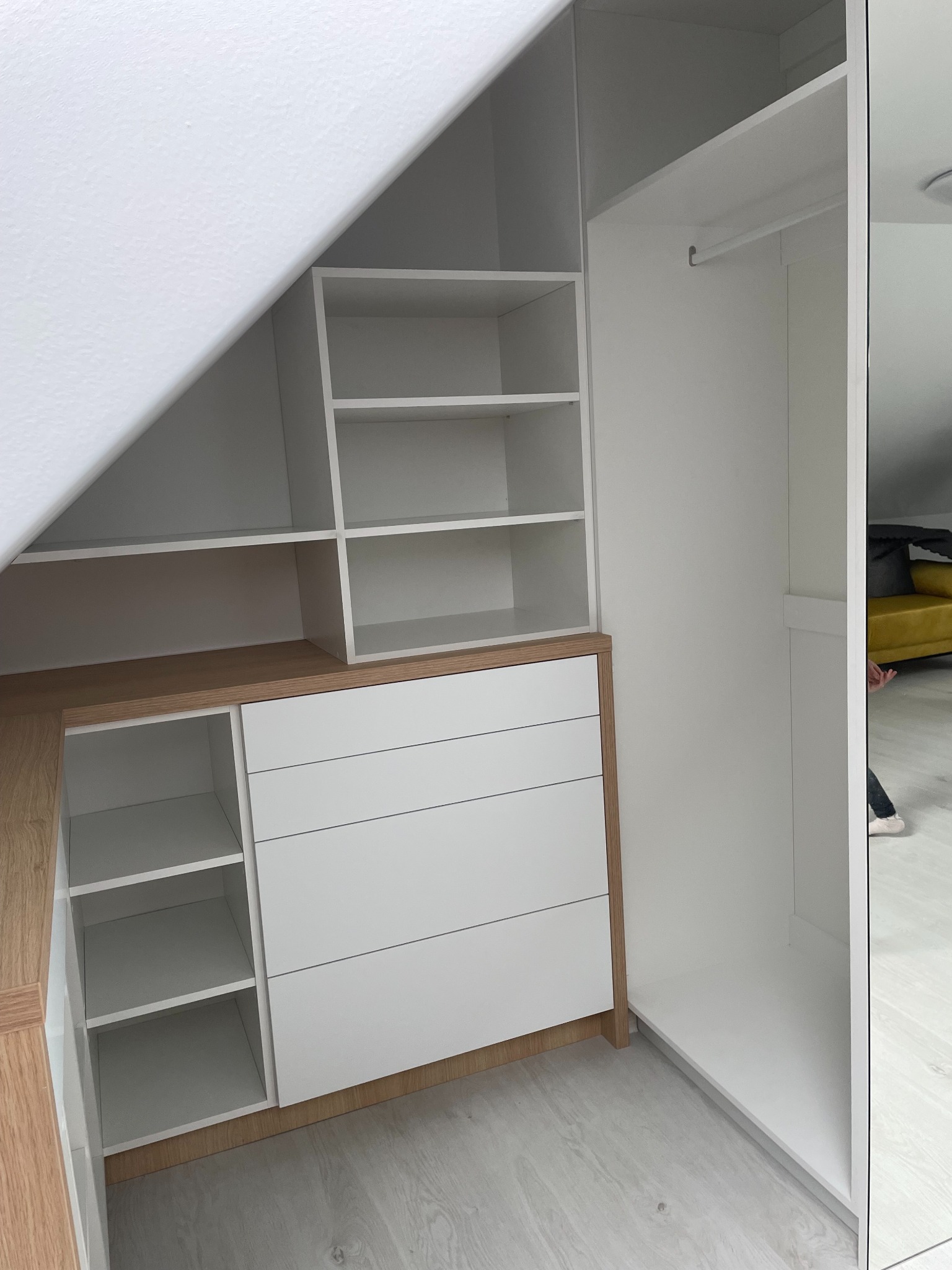 Biała szafa wnękowa z szufladami i półkami, dopasowana do skosu dachu, z drewnianym blatem. Widok z perspektywy wnętrza szafy, z fragmentem lustra po prawej.