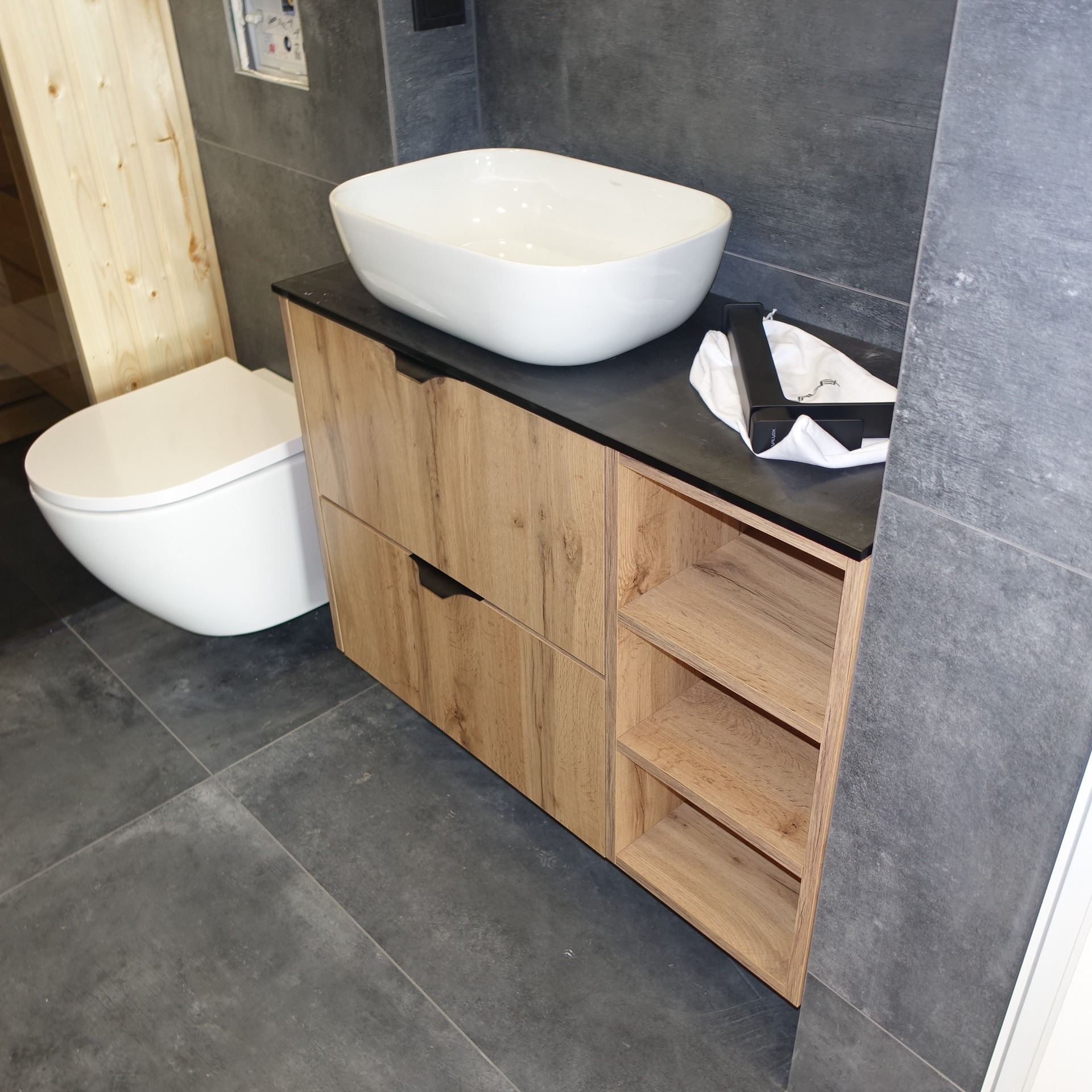 Drewniana szafka pod umywalkę z półkami i szufladami, czarny blat, biała umywalka nablatowa. Nowoczesna łazienka w Nowym Sączu.