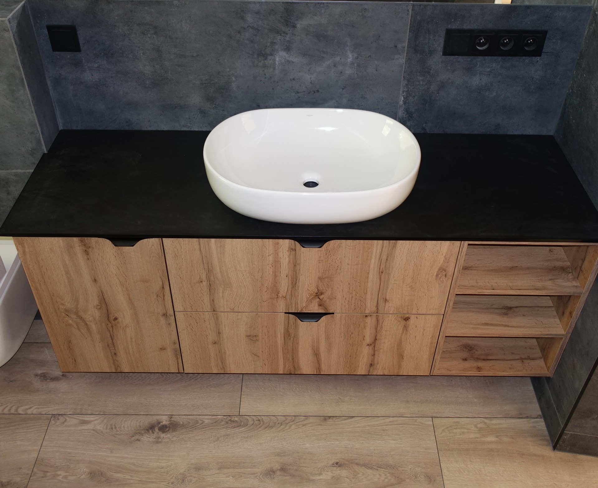 Nowoczesna szafka łazienkowa z blatem w kolorze czarnym i umywalką nablatową w kształcie owalnym. Fronty szafki w odcieniach drewna. Widoczne półki po prawej stronie.