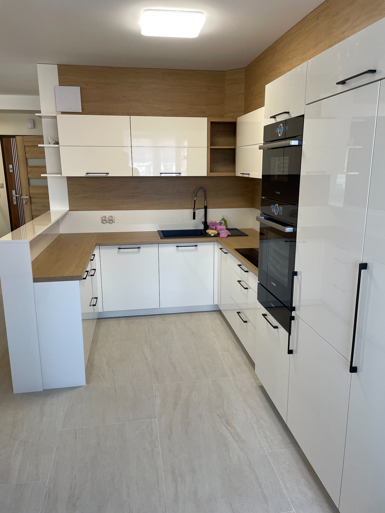 Nowoczesna kuchnia z białymi, lakierowanymi frontami i drewnianym blatem. W zabudowie piekarniki i płyta indukcyjna. Minimalistyczne, czarne uchwyty.