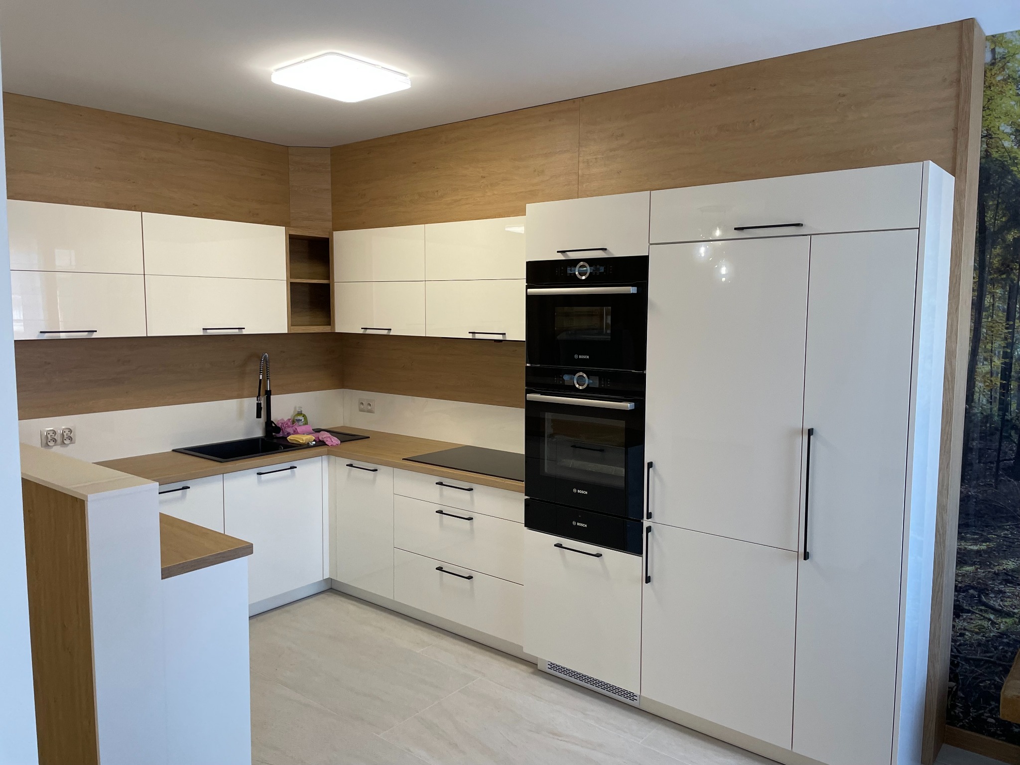 Nowoczesna kuchnia na wymiar z białymi, lakierowanymi frontami, czarnymi uchwytami i sprzętem AGD marki Bosch, z drewnianym panelem ściennym w tle.