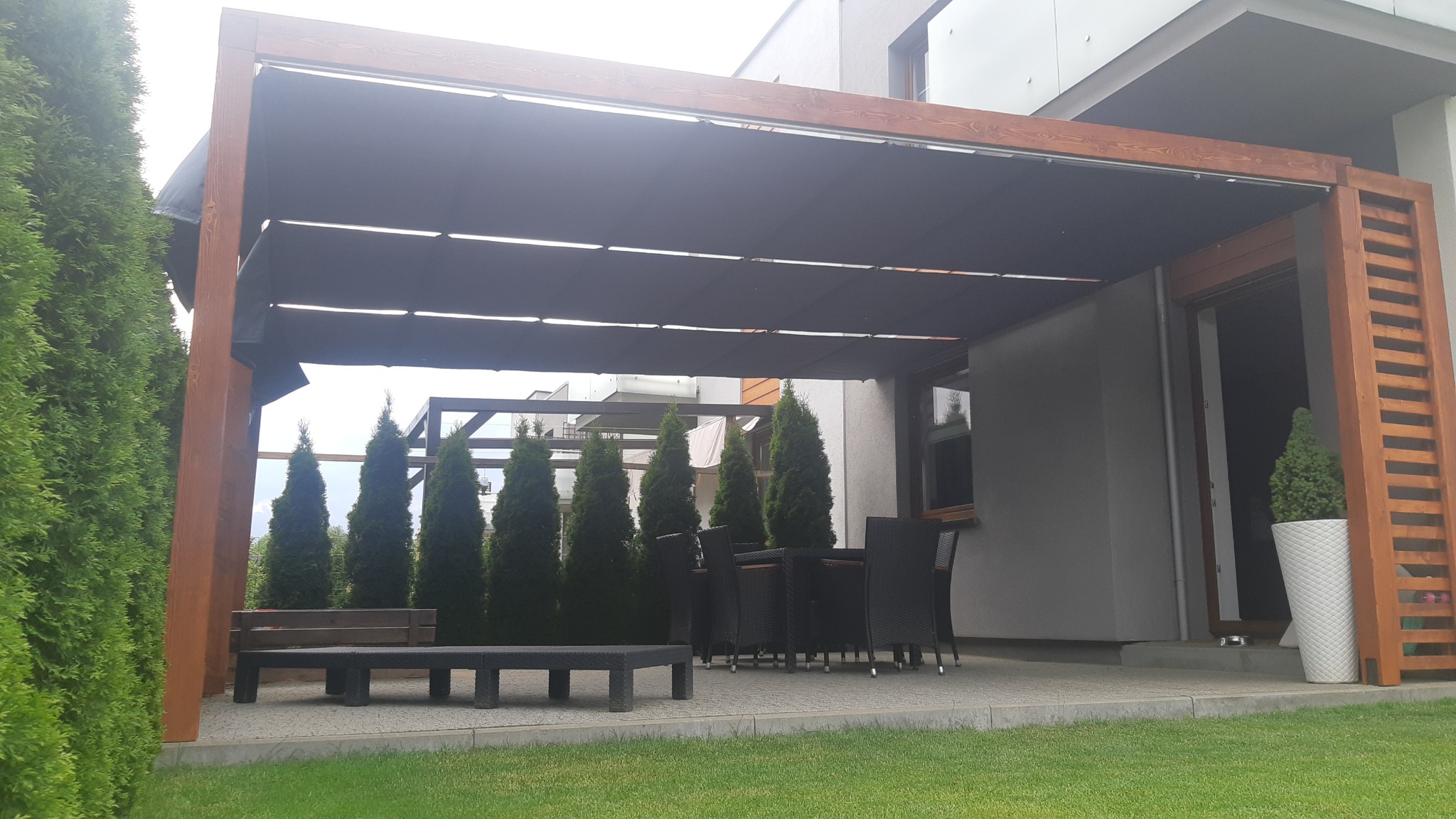 Drewniana konstrukcja z rozsuwanym, zaciemniającym dachem nad tarasem, widok na zestaw mebli ogrodowych i rząd strzelistych iglaków.