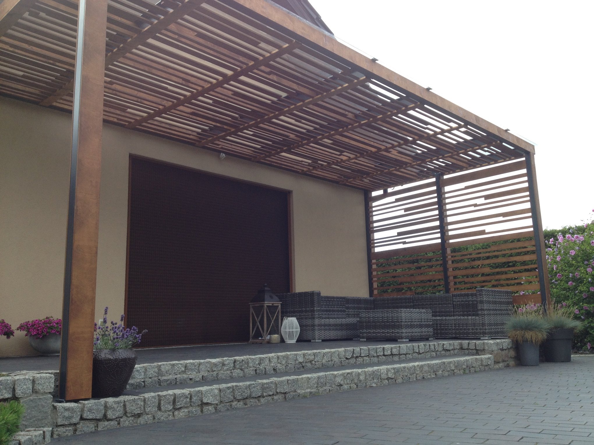 Drewniana pergola z poziomymi belkami w Poznaniu, z szarą sofą ogrodową i dekoracjami, na tle ściany budynku i zieleni.