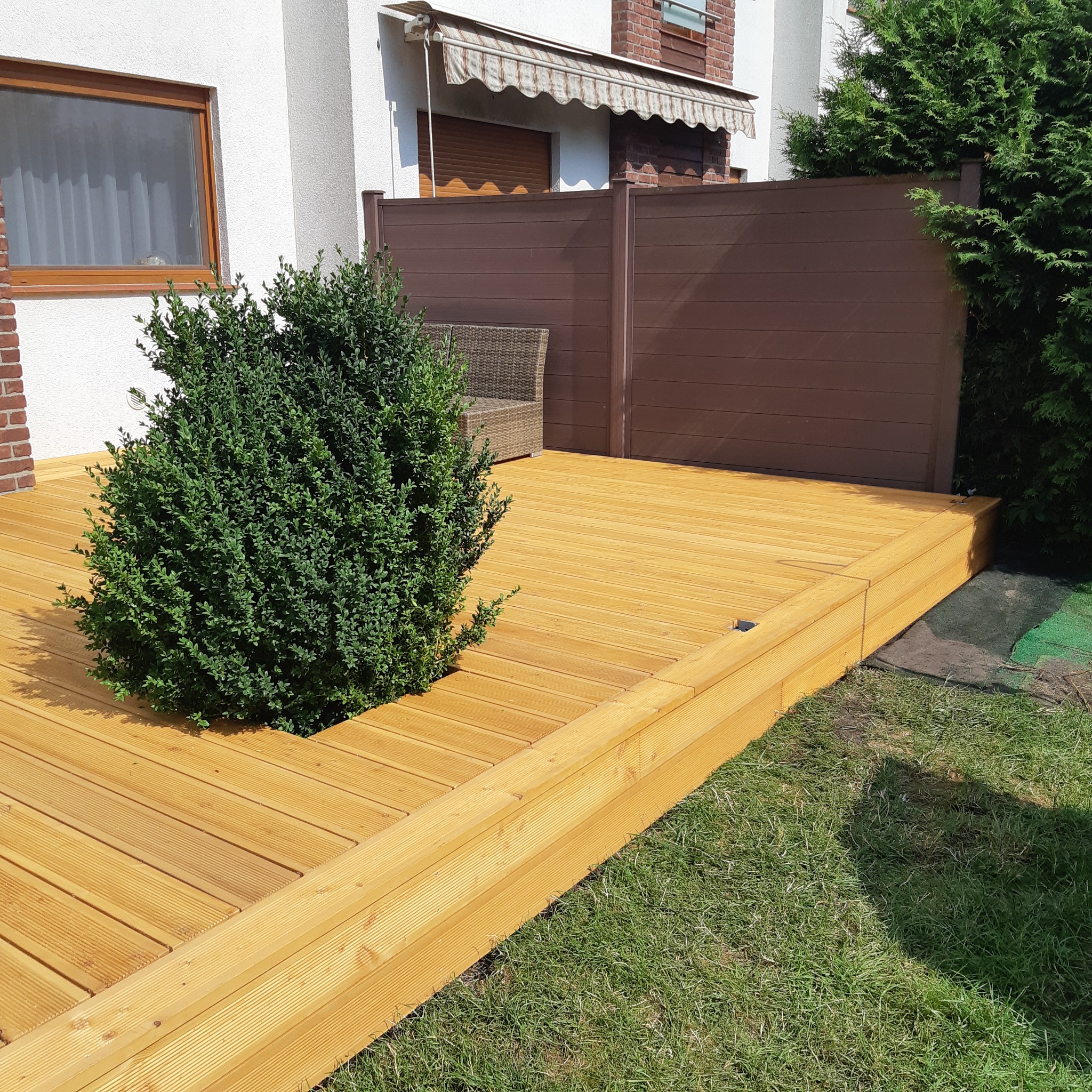 Jasny, drewniany taras z wbudowaną rabatą, w której rośnie gęsty, kulisty krzew. W tle widoczny fragment domu z oknem i brązowe ogrodzenie.