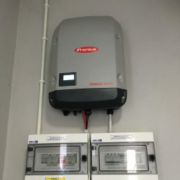 KMBSOLAR SPÓŁKA Z OGRANICZONĄ ODPOWIEDZIALNOŚCIĄ - Szary falownik Fronius Symo zamontowany na ścianie nad dwiema białymi rozdzielnicami PV DC i PV AC. Widoczne przewody i rury instalacyjne. Na wyświetlaczu falownika widoczny numer.