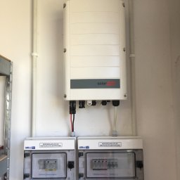 KMBSOLAR SPÓŁKA Z OGRANICZONĄ ODPOWIEDZIALNOŚCIĄ - Biały inwerter SolarEdge zamontowany na ścianie nad dwiema rozdzielnicami elektrycznymi PV DC i PV AC w pomieszczeniu technicznym.