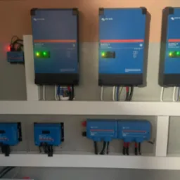 Instalacja z trzema falownikami MultiPlus-II Victron Energy, kontrolerami ładowania MPPT i modułami Lynx Power In, zamontowana na ścianie wewnątrz pomieszczenia. Widoczne przewody i diody LED...