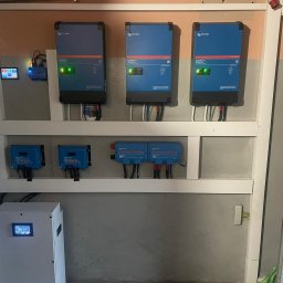 KMBSOLAR SPÓŁKA Z OGRANICZONĄ ODPOWIEDZIALNOŚCIĄ - Instalacja z trzema niebieskimi falownikami MultiPlus-II, dwoma regulatorami ładowania MPPT i magazynem energii na ścianie w pomieszczeniu technicznym.