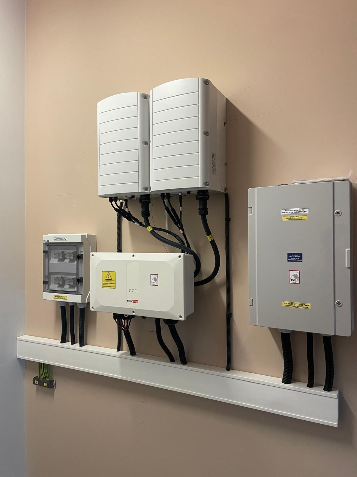 Białe skrzynki z okablowaniem, prawdopodobnie inwertery, zamontowane na ścianie wewnątrz budynku, z widocznymi oznaczeniami i tabliczkami ostrzegawczymi.