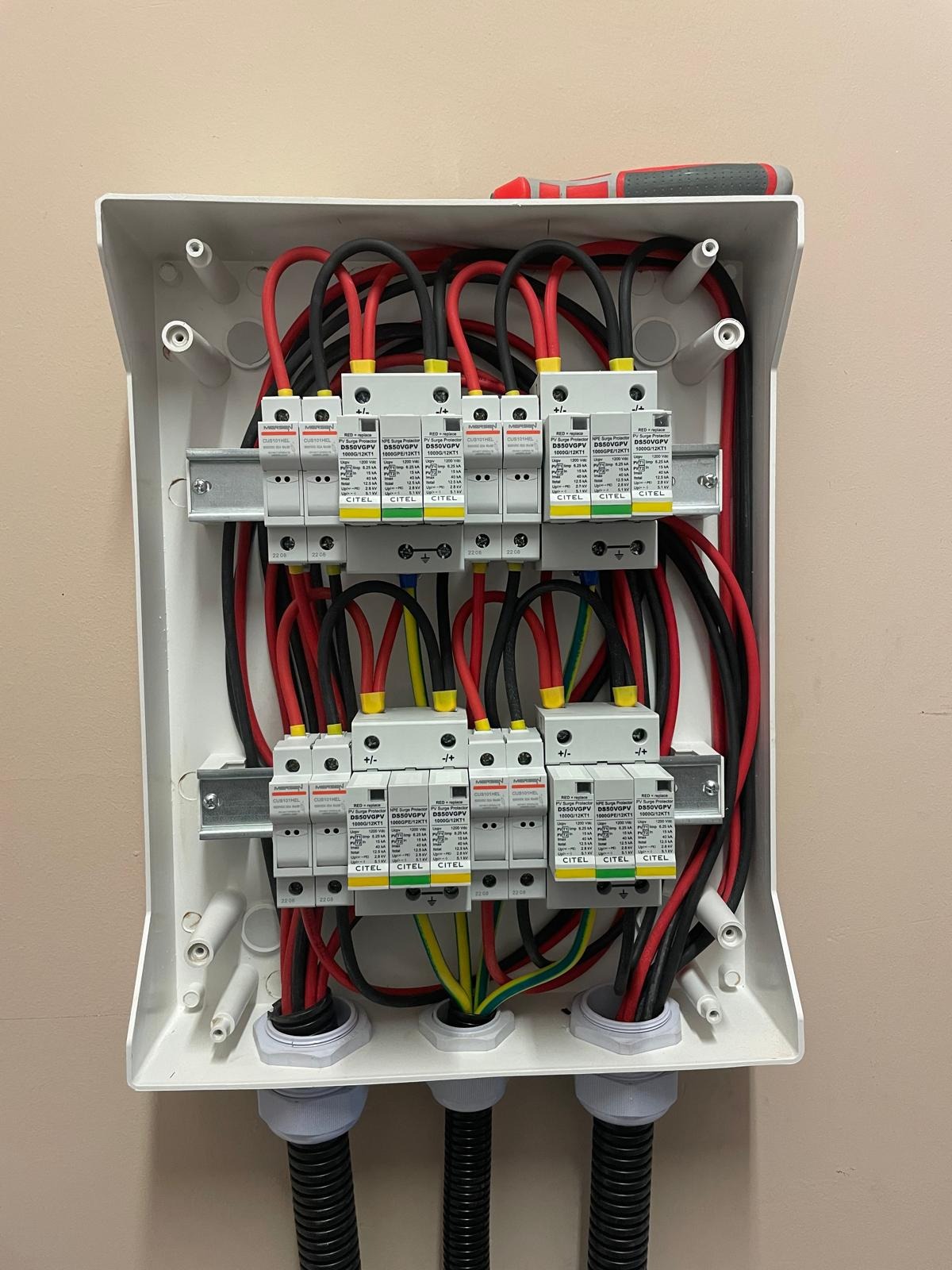 Rozdzielnica elektryczna z zabezpieczeniami przeciwprzepięciowymi, widoczne przewody w kolorach czarnym i czerwonym oraz żółto-zielonym, ułożone wewnątrz białej obudowy, na górze obudowy leży...