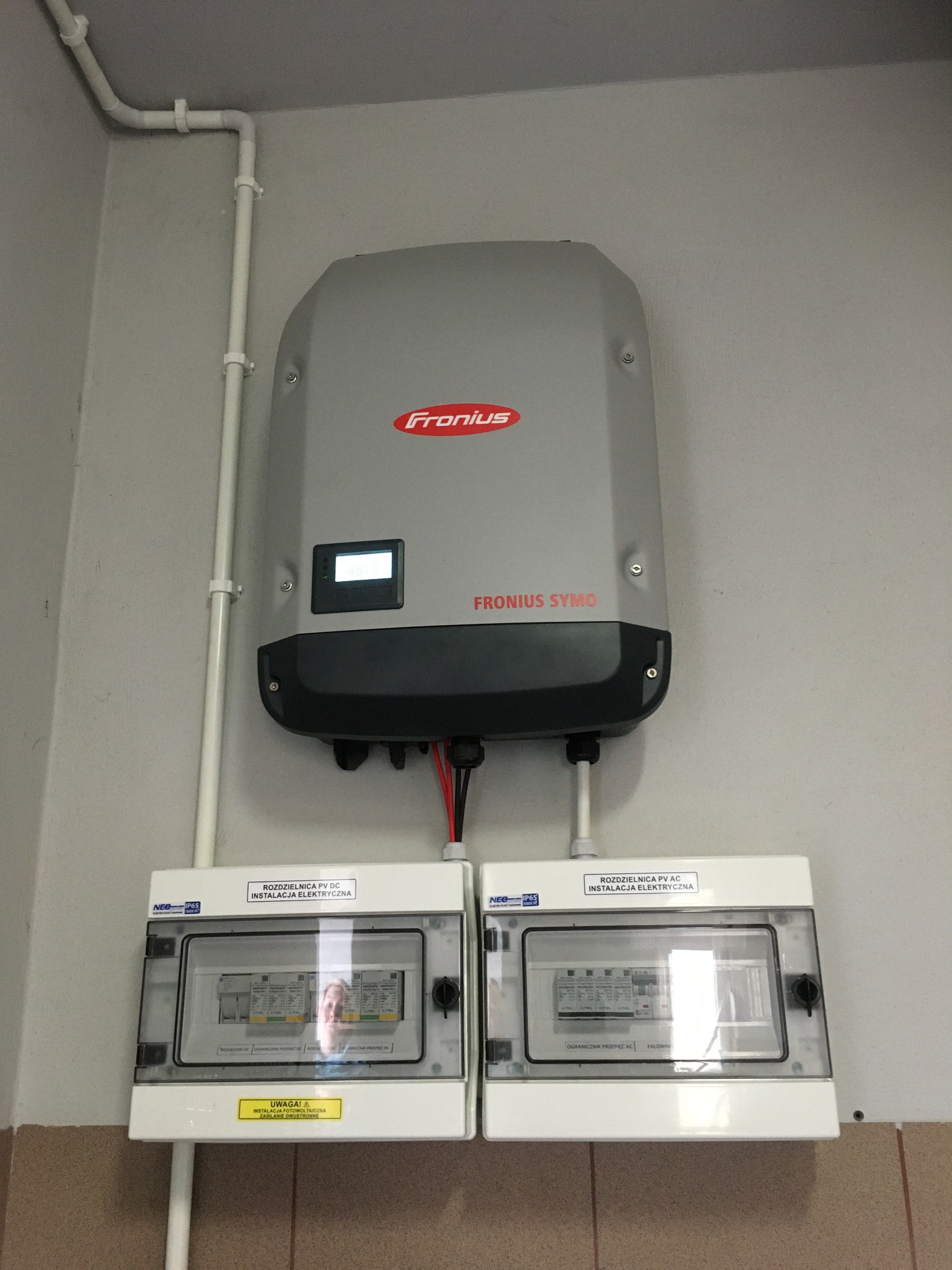 Szary falownik Fronius Symo zamontowany na ścianie nad dwiema białymi rozdzielnicami PV DC i PV AC. Widoczne przewody i rury instalacyjne. Na wyświetlaczu falownika widoczny numer.