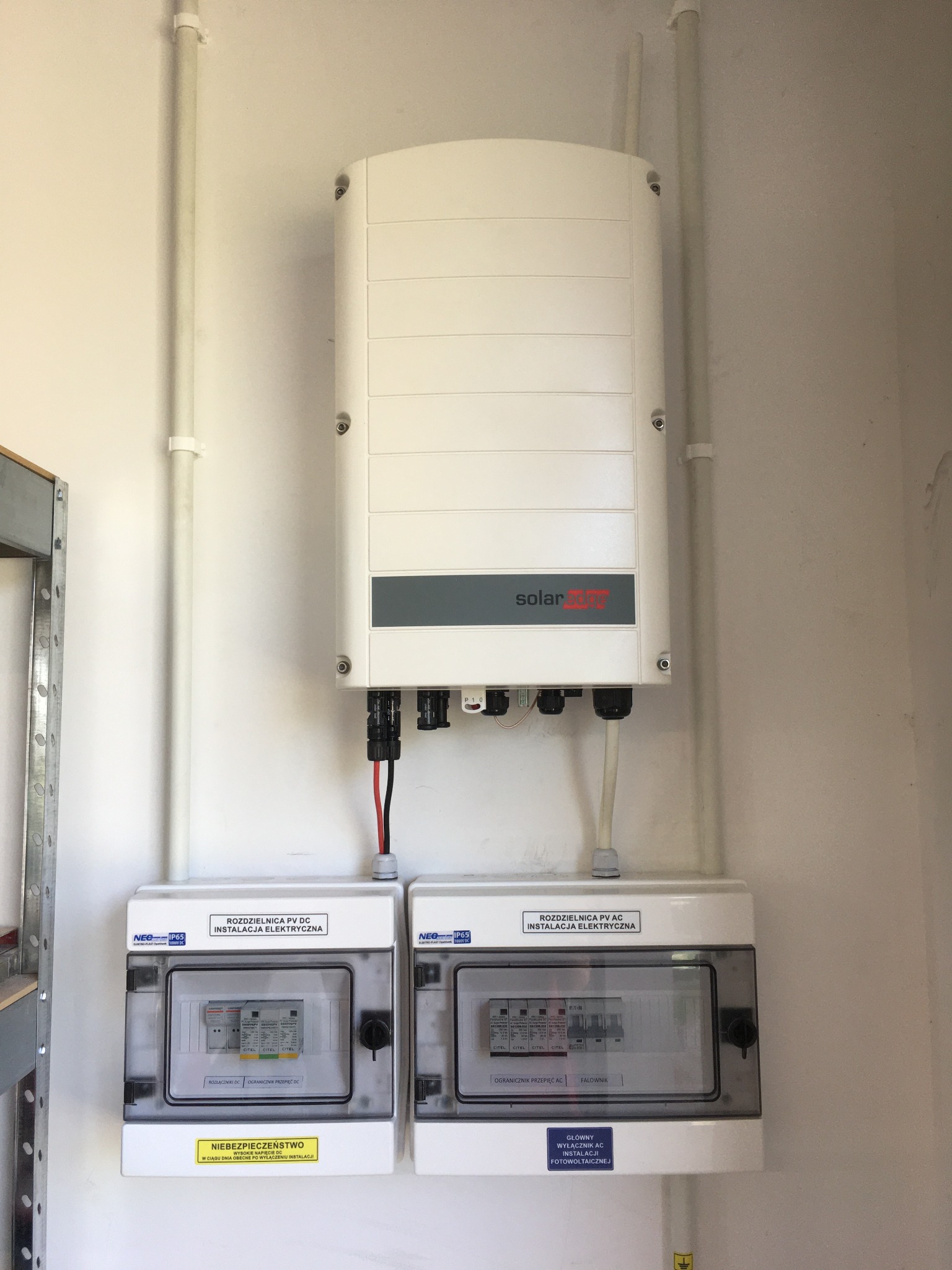 Biały inwerter SolarEdge zamontowany na ścianie nad dwiema rozdzielnicami elektrycznymi PV DC i PV AC w pomieszczeniu technicznym.