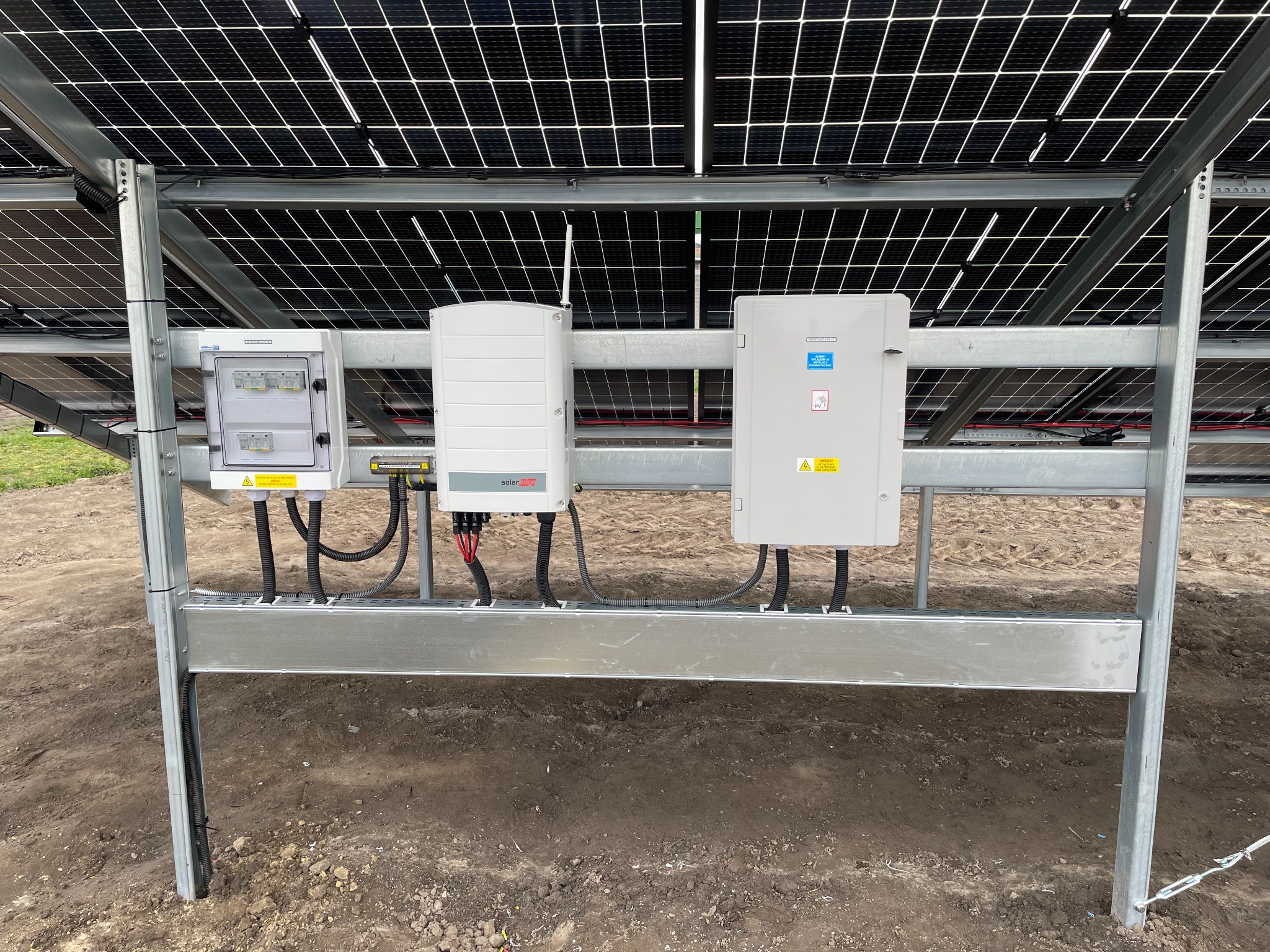 Instalacja paneli słonecznych z widoczną skrzynką elektryczną, inwerterem SolarEdge i zabezpieczeniami, zamontowanymi na metalowej konstrukcji, na tle ziemi.