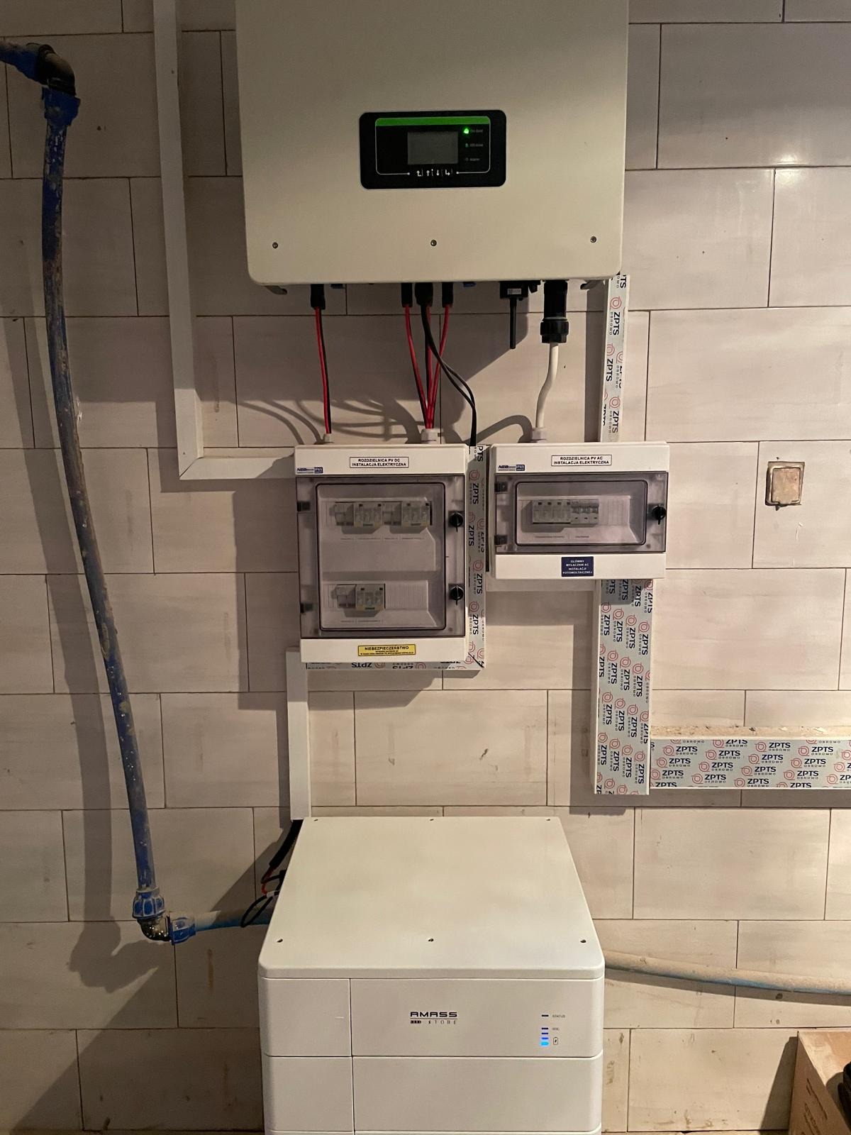Kompletna instalacja domowa: falownik, rozdzielnice elektryczne i akumulator energii AMASS, połączone przewodami w pomieszczeniu z kafelkowymi ścianami.