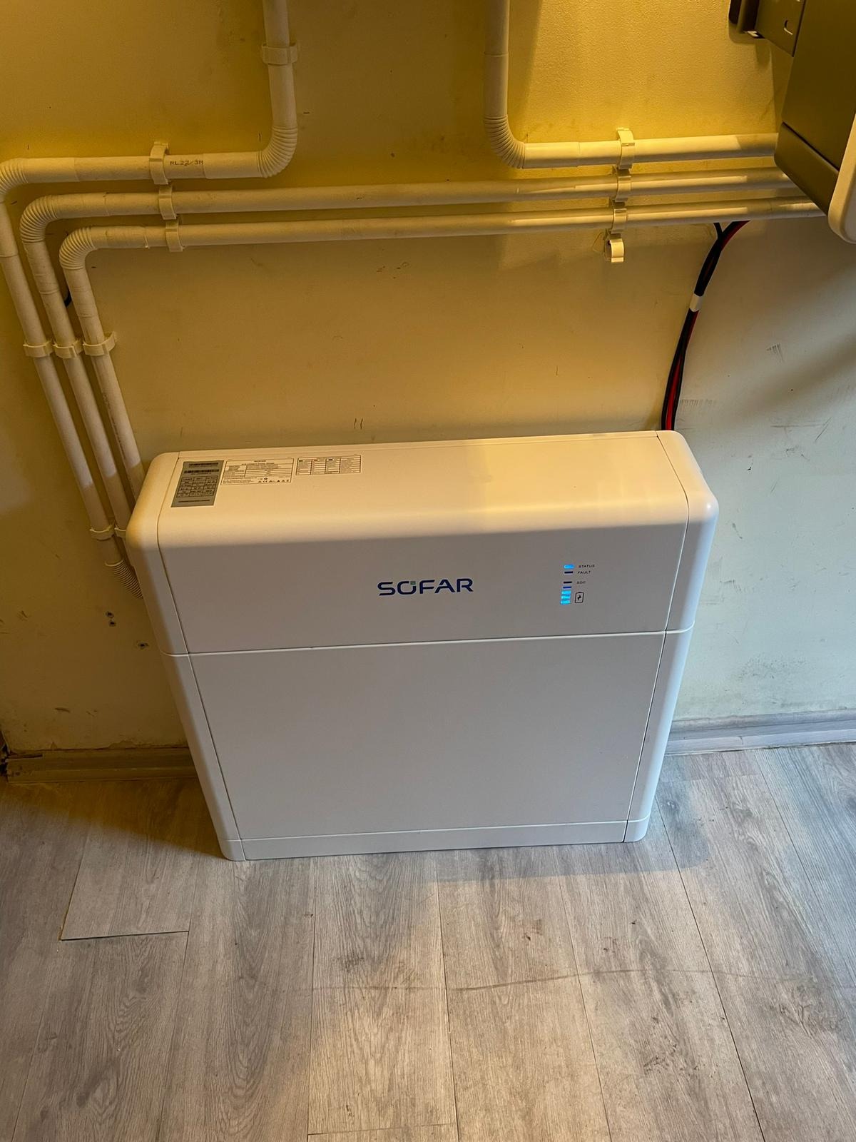 Biały magazyn energii Sofar umieszczony przy żółtej ścianie z instalacją rur, widoczne niebieskie diody statusu i podłoga imitująca drewno.