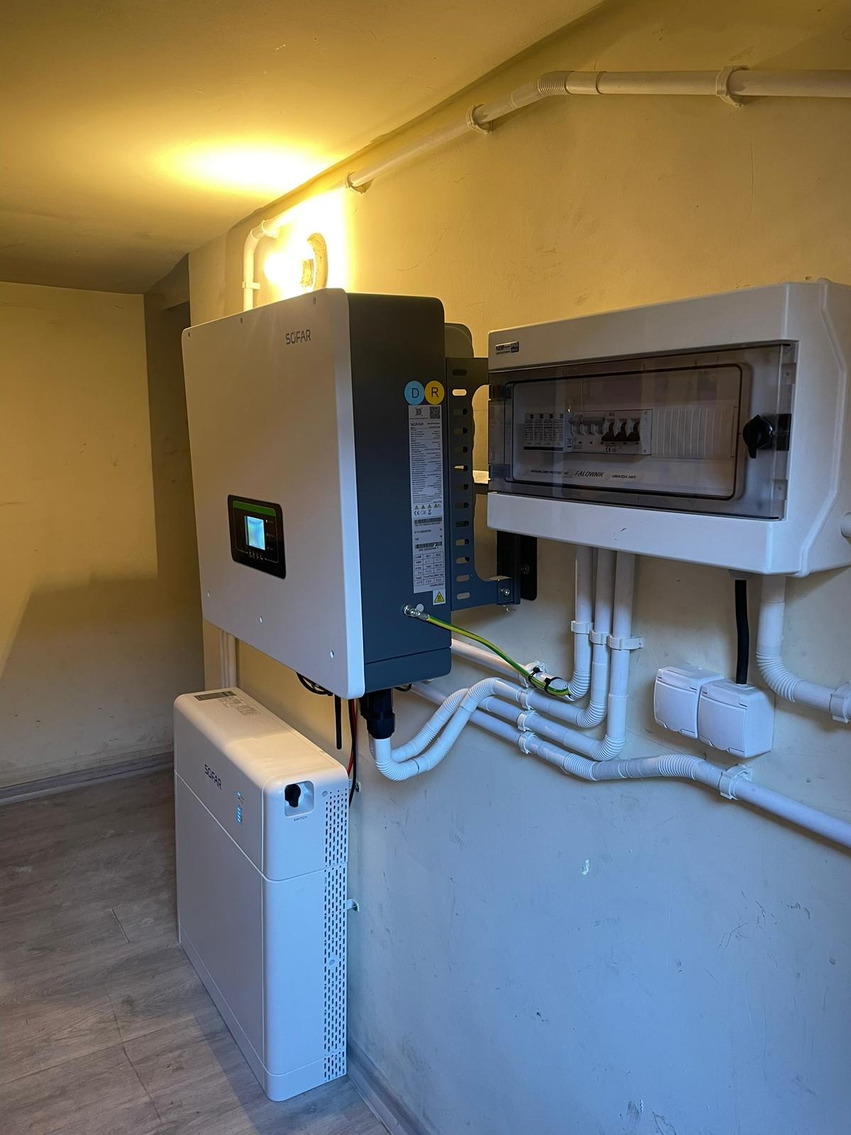 Instalacja wewnętrzna: falownik SOFAR, magazyn energii oraz skrzynka z bezpiecznikami, widoczne przewody elektryczne w białych peszlach