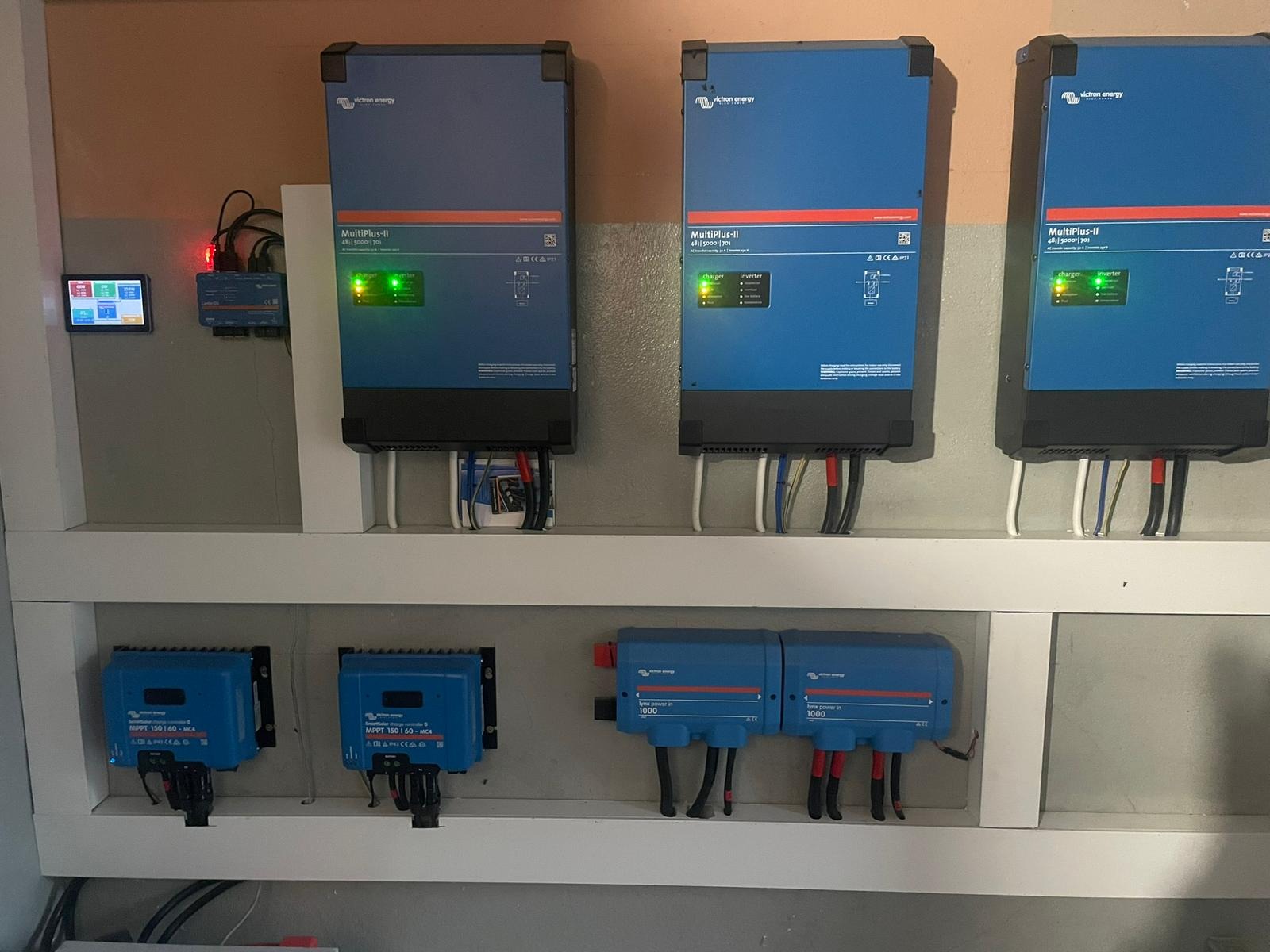Instalacja z trzema falownikami MultiPlus-II Victron Energy, kontrolerami ładowania MPPT i modułami Lynx Power In, zamontowana na ścianie wewnątrz pomieszczenia. Widoczne przewody i diody LED...