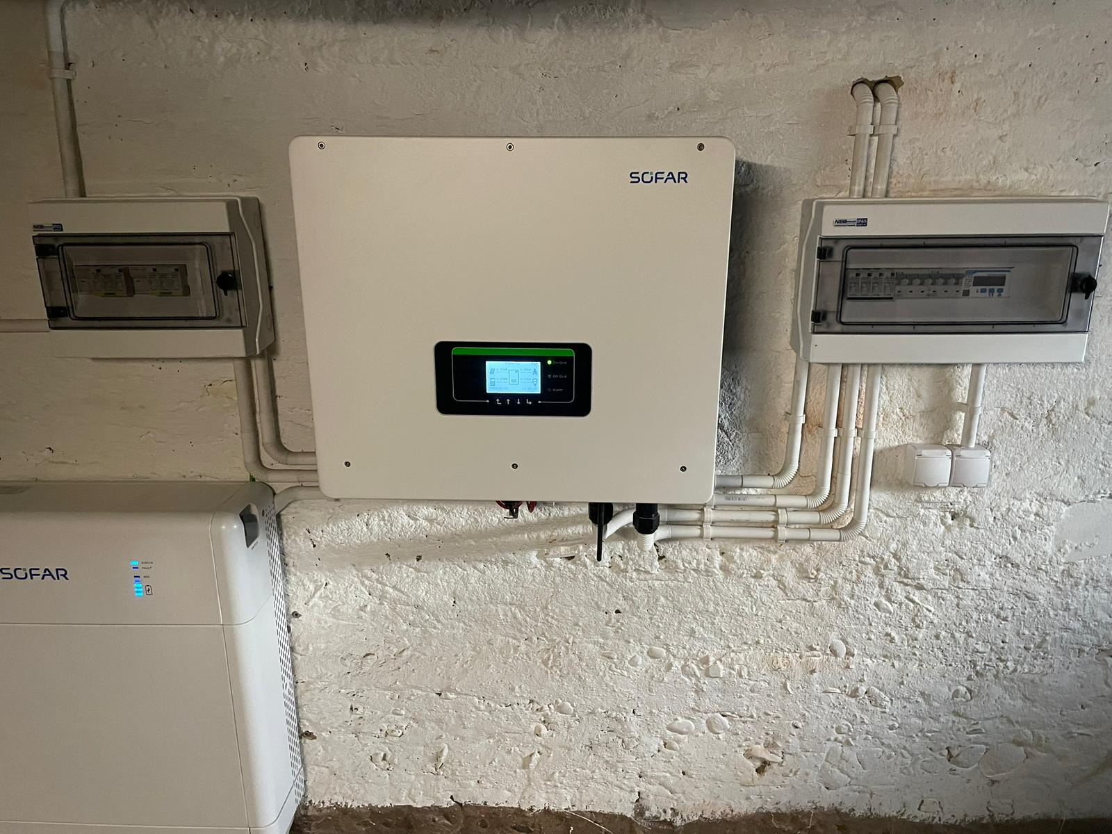 Biały inwerter SOFAR z wyświetlaczem i biały akumulator energii SOFAR zamontowane na nierównej, pomalowanej na biało ścianie z widocznymi rurami i skrzynkami elektrycznymi.