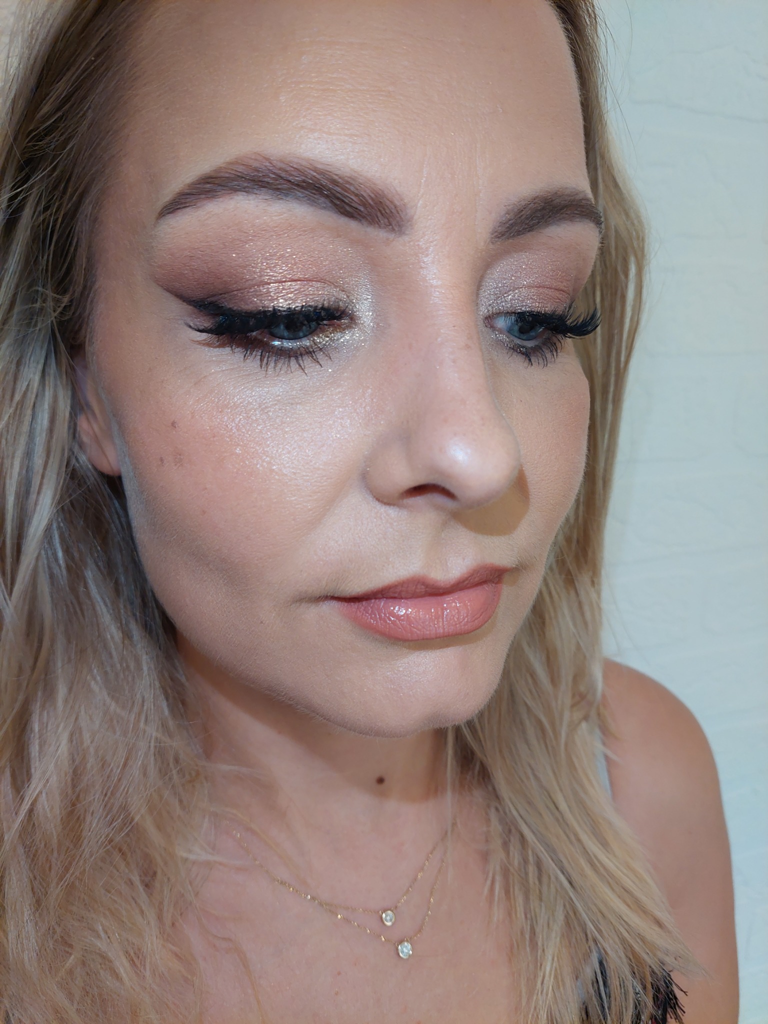 Portret kobiety z delikatnym, rozświetlonym cieniem na powiekach, precyzyjną kreską eyelinerem i błyszczącymi ustami w odcieniu nude, patrzącej w dół.