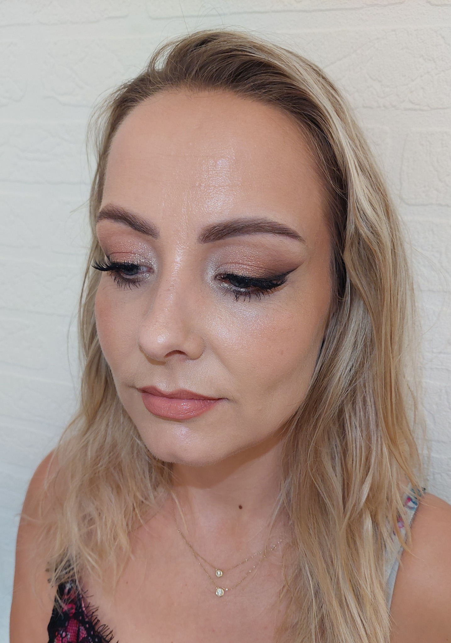 Portret kobiety z delikatnym, błyszczącym cieniem do powiek w odcieniach brązu i beżu, czarną kreską eyelinerem i błyszczącymi, naturalnymi ustami. Widoczna złota biżuteria na szyi.