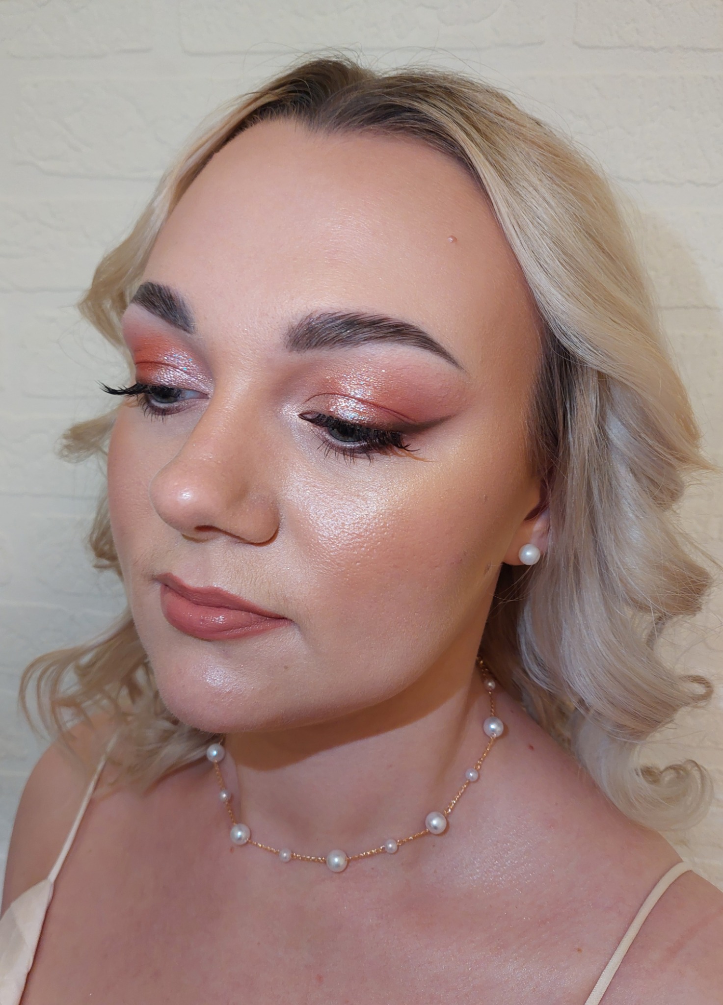 Portret kobiety z delikatnym, błyszczącym rumieńcem i pomarańczowo-różowym cieniem na powiekach, uzupełniony perłową biżuterią.