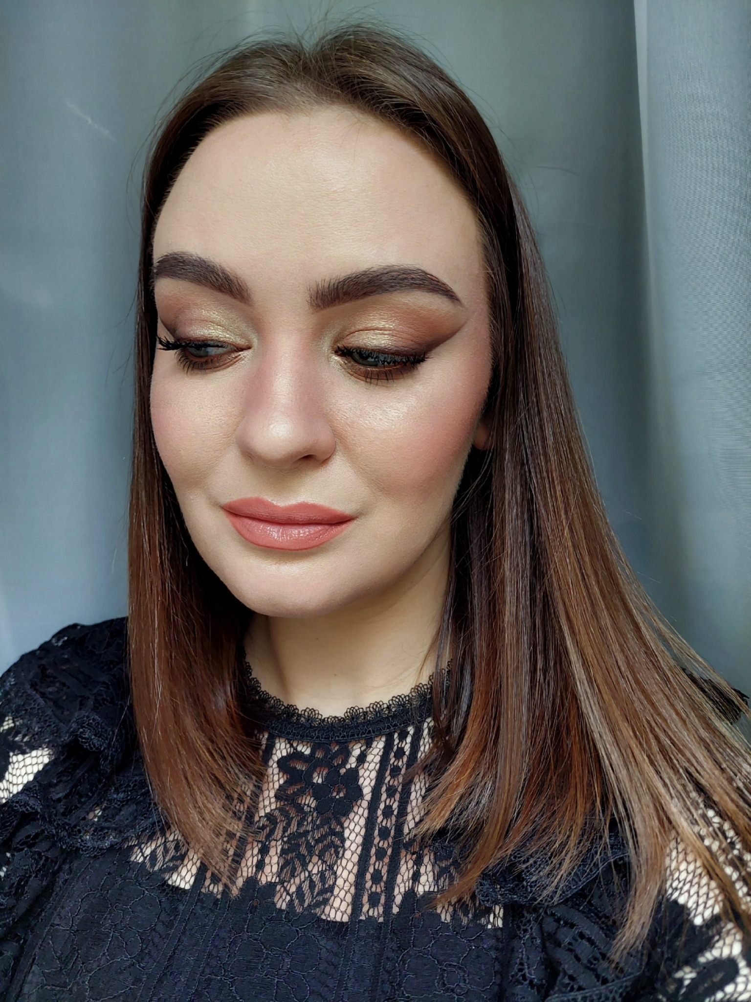 Portret kobiety z makijażem w odcieniach brązu i złota, z wyraźnie zaznaczonymi rzęsami i ustami w kolorze nude, ubrana w czarną koronkową bluzkę.