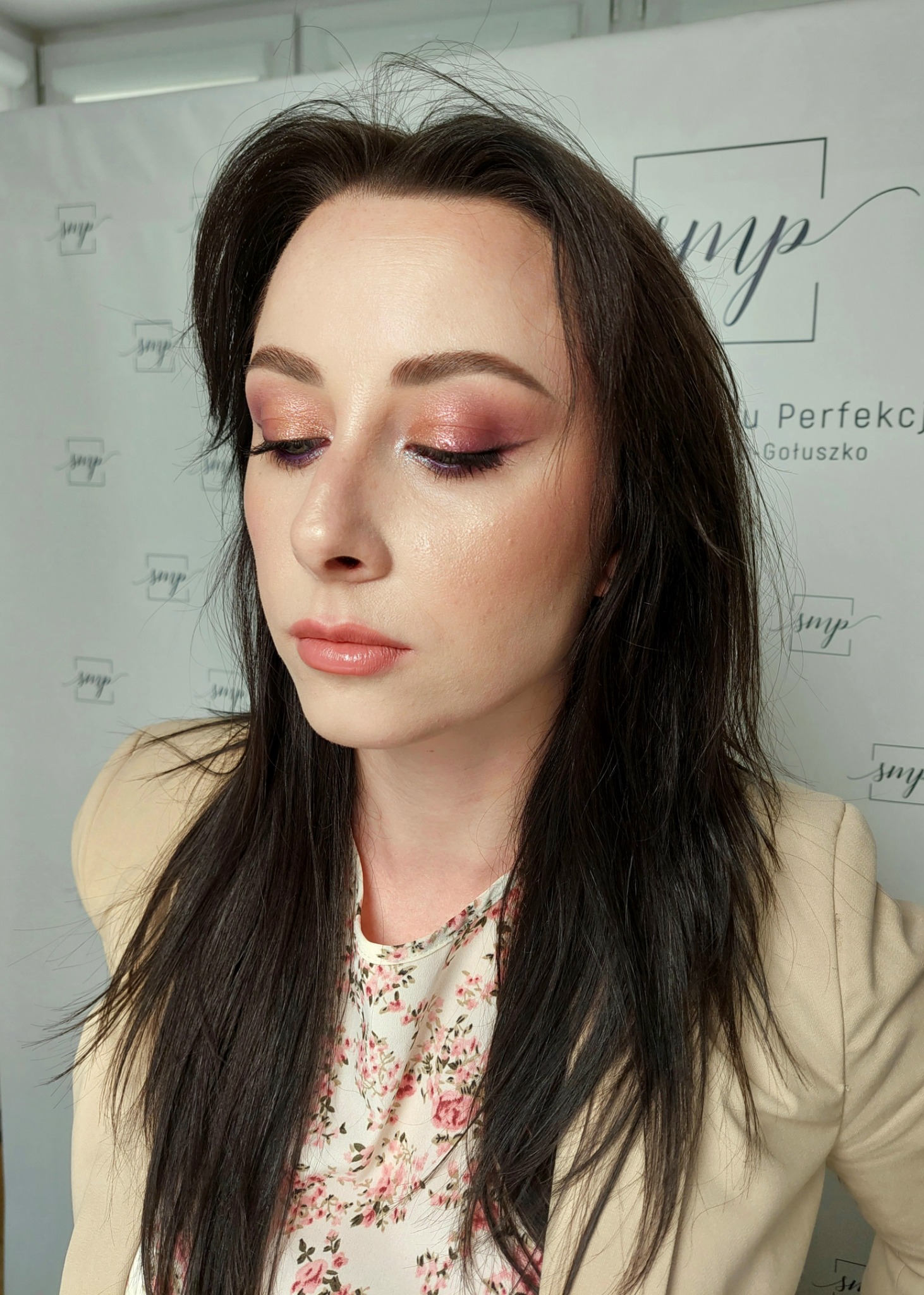 Portret kobiety z delikatnym, różowym makijażem oczu i ust. Świeża cera, ciemne włosy. Ujęcie z bliska, w tle logo firmy kosmetycznej.