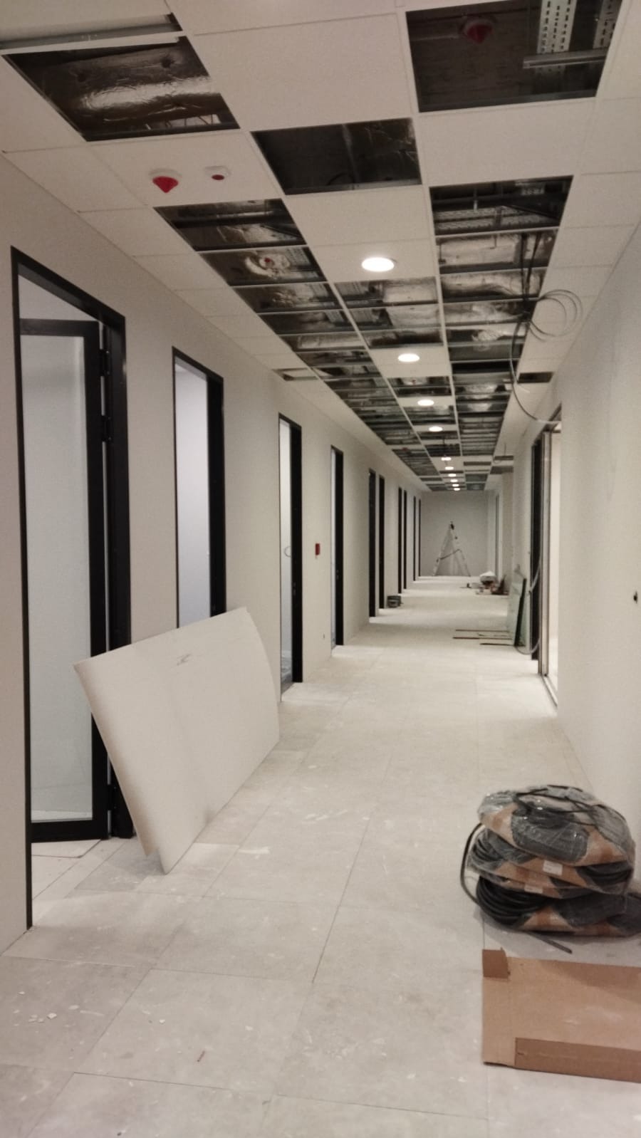 Korytarz biurowy w trakcie wykańczania: widoczne otwarte sufity podwieszane z instalacjami, przygotowane płyty sufitowe oparte o ścianę, oraz zwój kabli na podłodze.