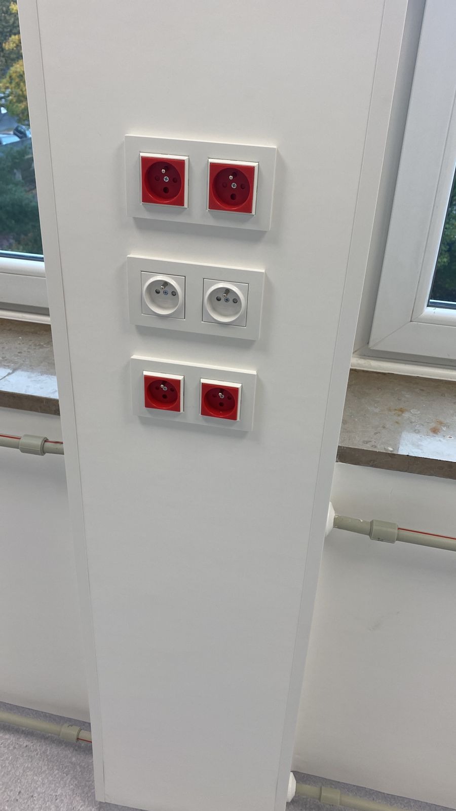 Biała ściana z trzema rzędami podwójnych gniazdek elektrycznych: dwa rzędy z czerwonymi gniazdkami i jeden rząd z białymi gniazdkami, widoczne rury instalacyjne w tle, naturalne światło z okna.