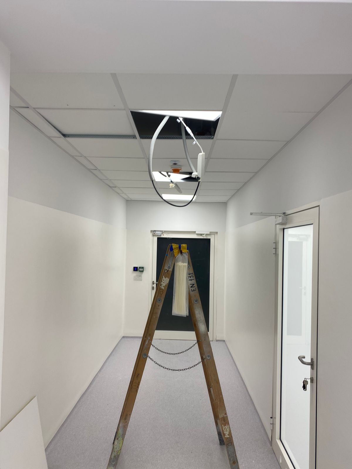 Korytarz w trakcie prac instalacyjnych: drabina oparta o drzwi, widoczne przewody wystające z otworu w suficie podwieszanym.