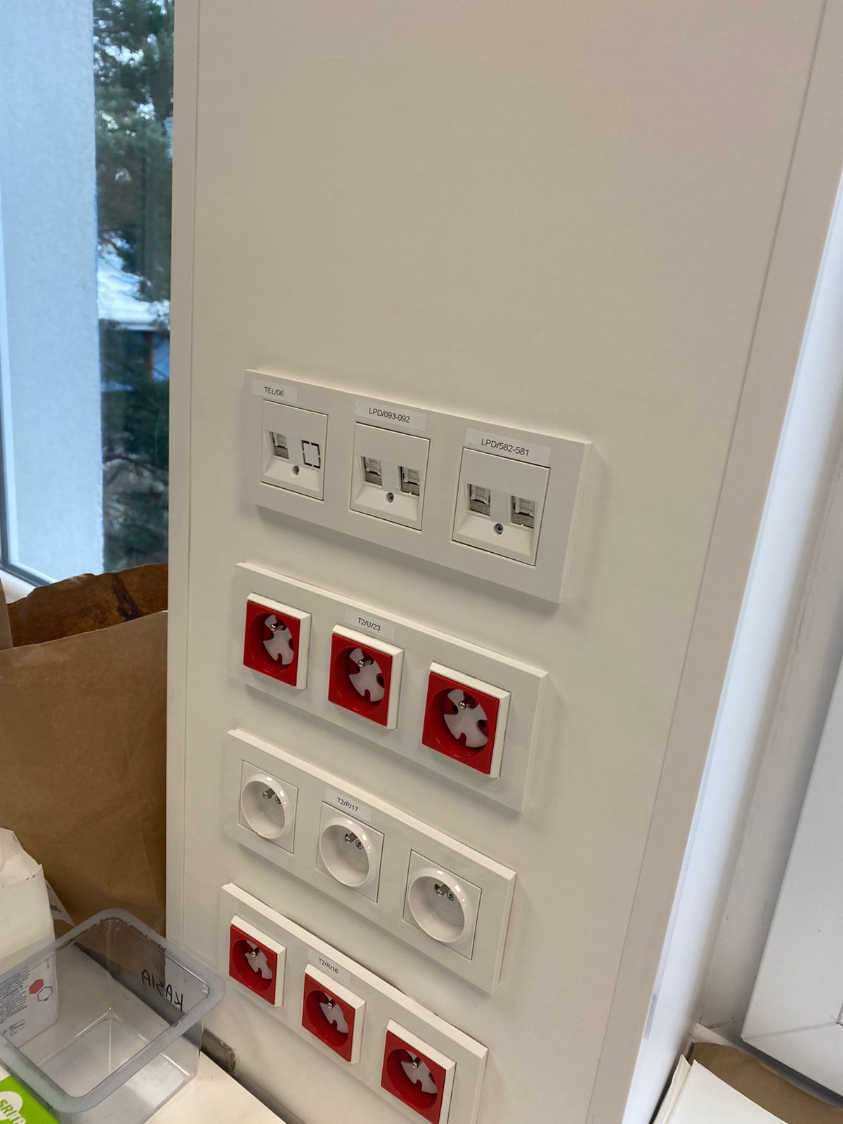 Rząd gniazd sieciowych, telefonicznych i alarmowych w białych ramkach zamontowanych na ścianie, widoczne napisy identyfikacyjne na obudowach, w tle okno i kartony.