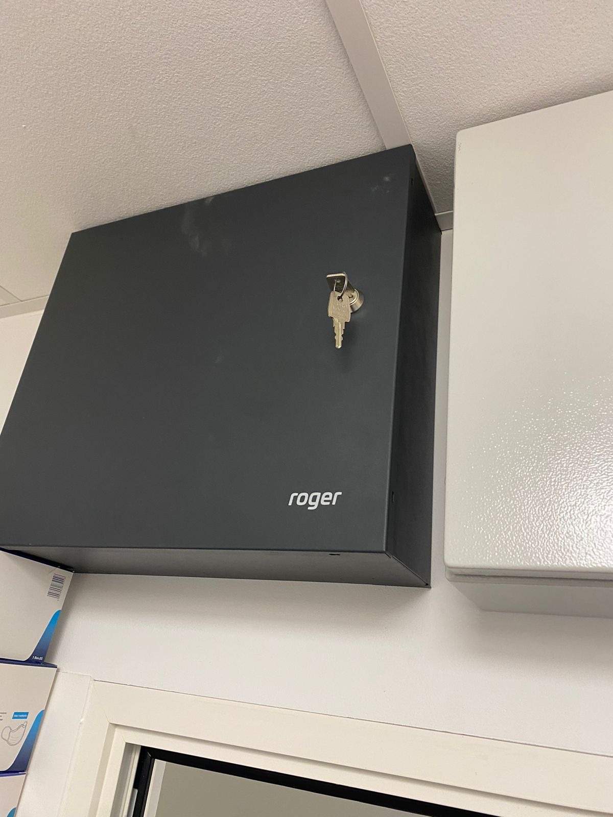 Szara szafka sterownicza firmy Roger zamontowana na ścianie, z kluczem w zamku, obok białej szafki, widok z dołu.